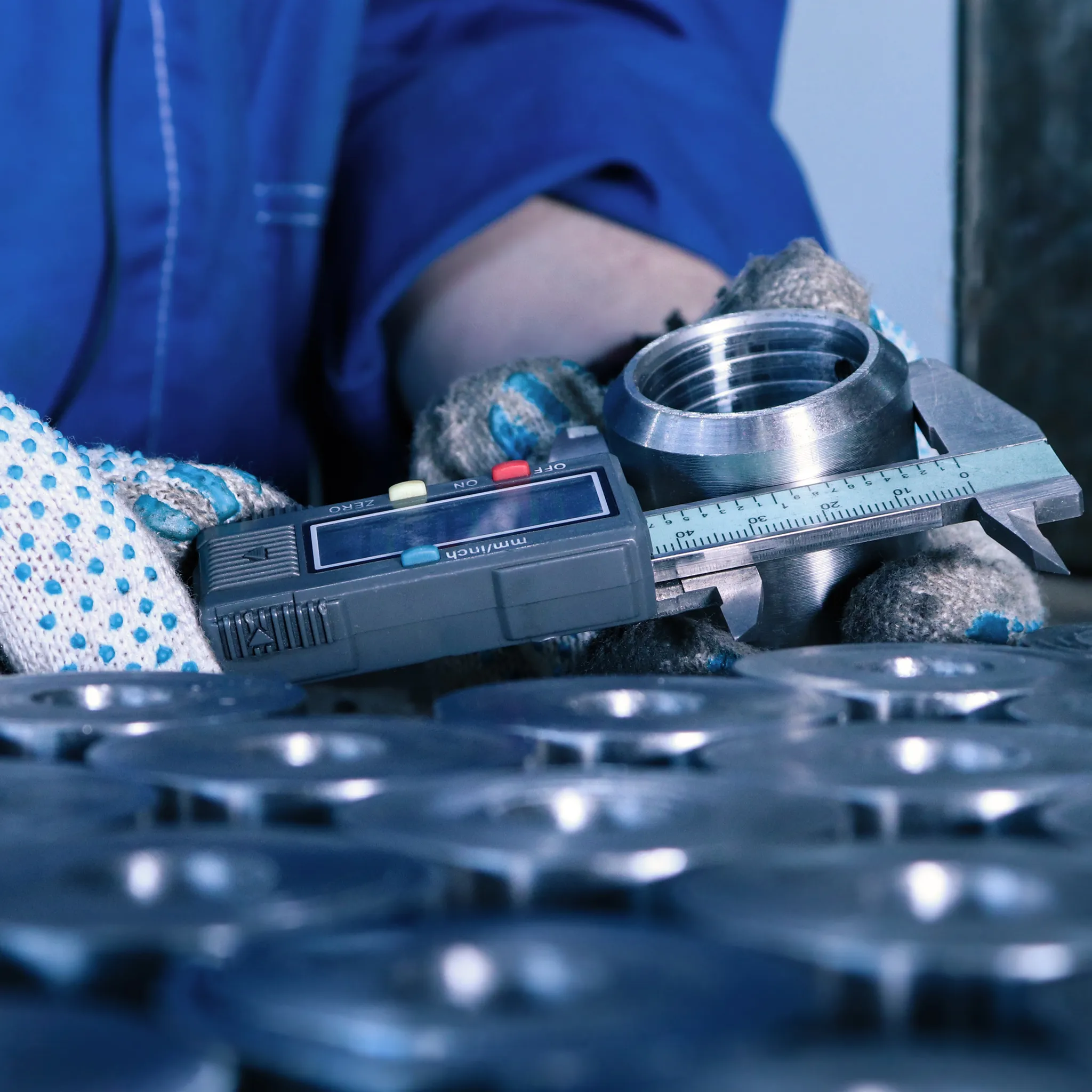 Ein Arbeiter in blauer Uniform und gepunkteten Handschuhen misst ein Metallteil mit einem digitalen Messschieber.