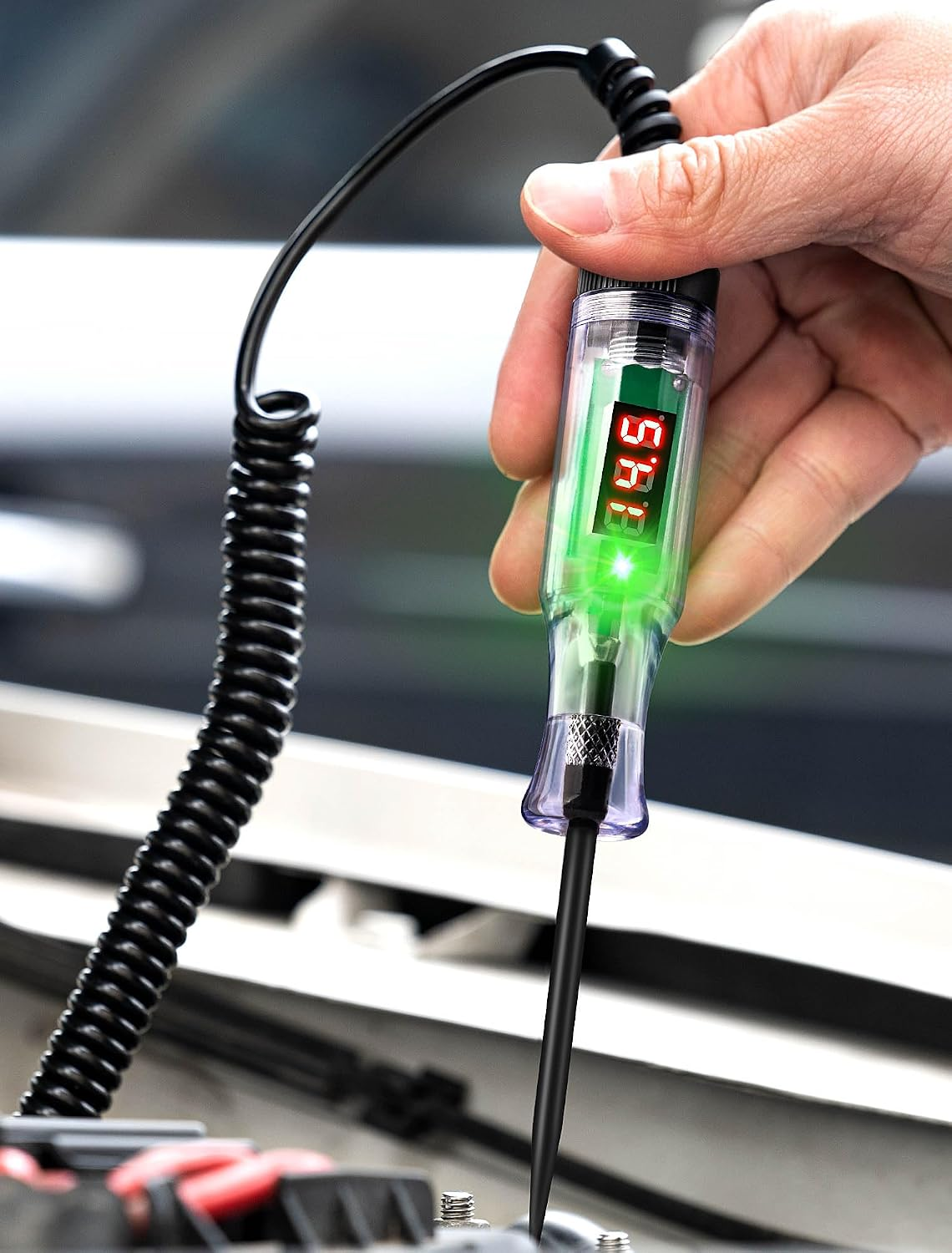 Hand hält einen digitalen Kfz-Spannungsprüfer mit grünem Licht und schwarzem Spiralkabel über einem Motorraum.