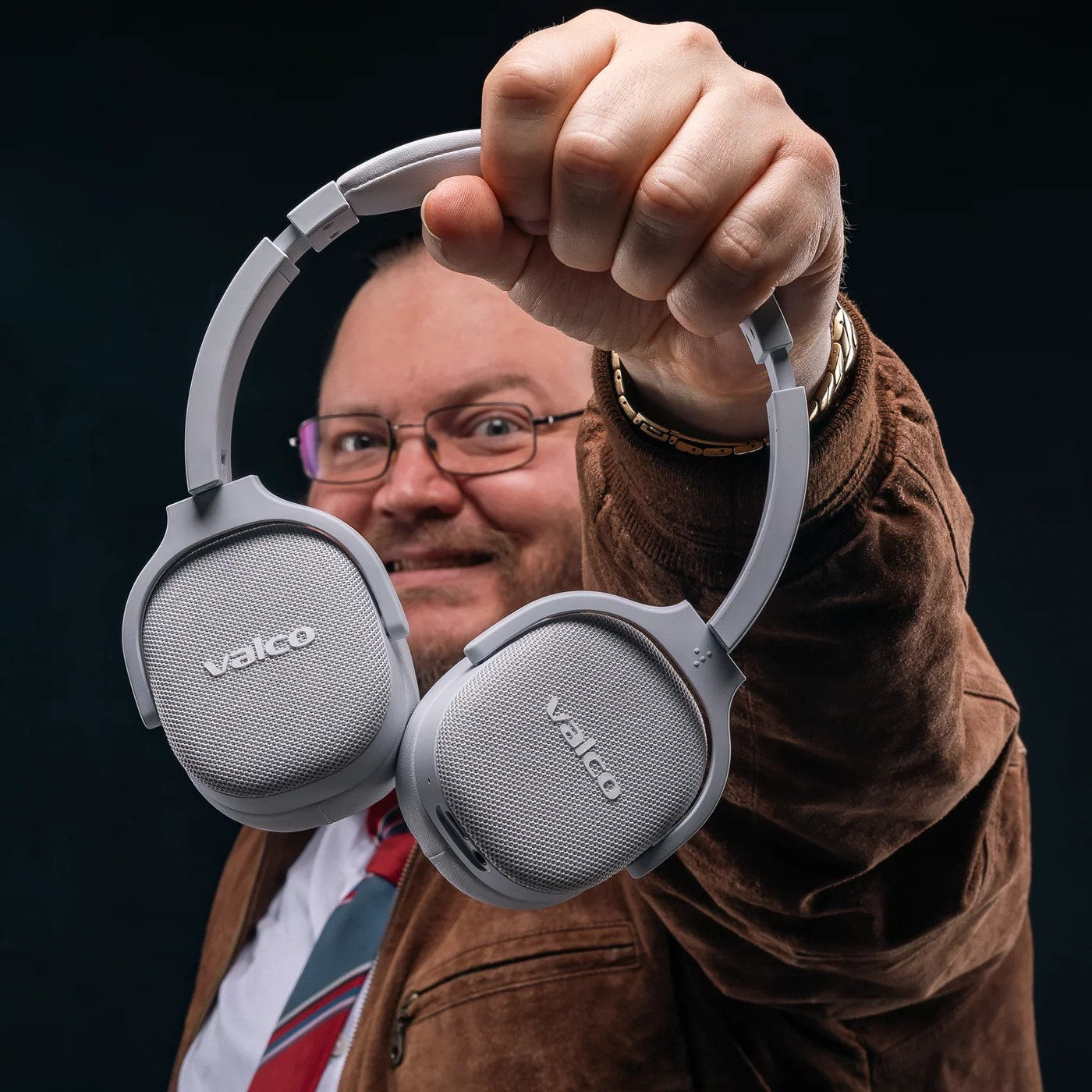 Ein Mann mit Brille und brauner Jacke hält graue Valco-Kopfhörer in die Kamera.