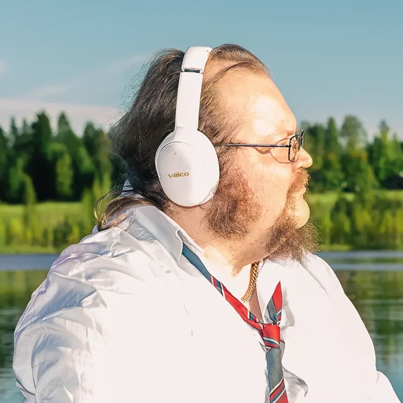 Mann mit Brille und weißen Kopfhörern, weißem Hemd und Krawatte vor einer Wasser- und Waldlandschaft.