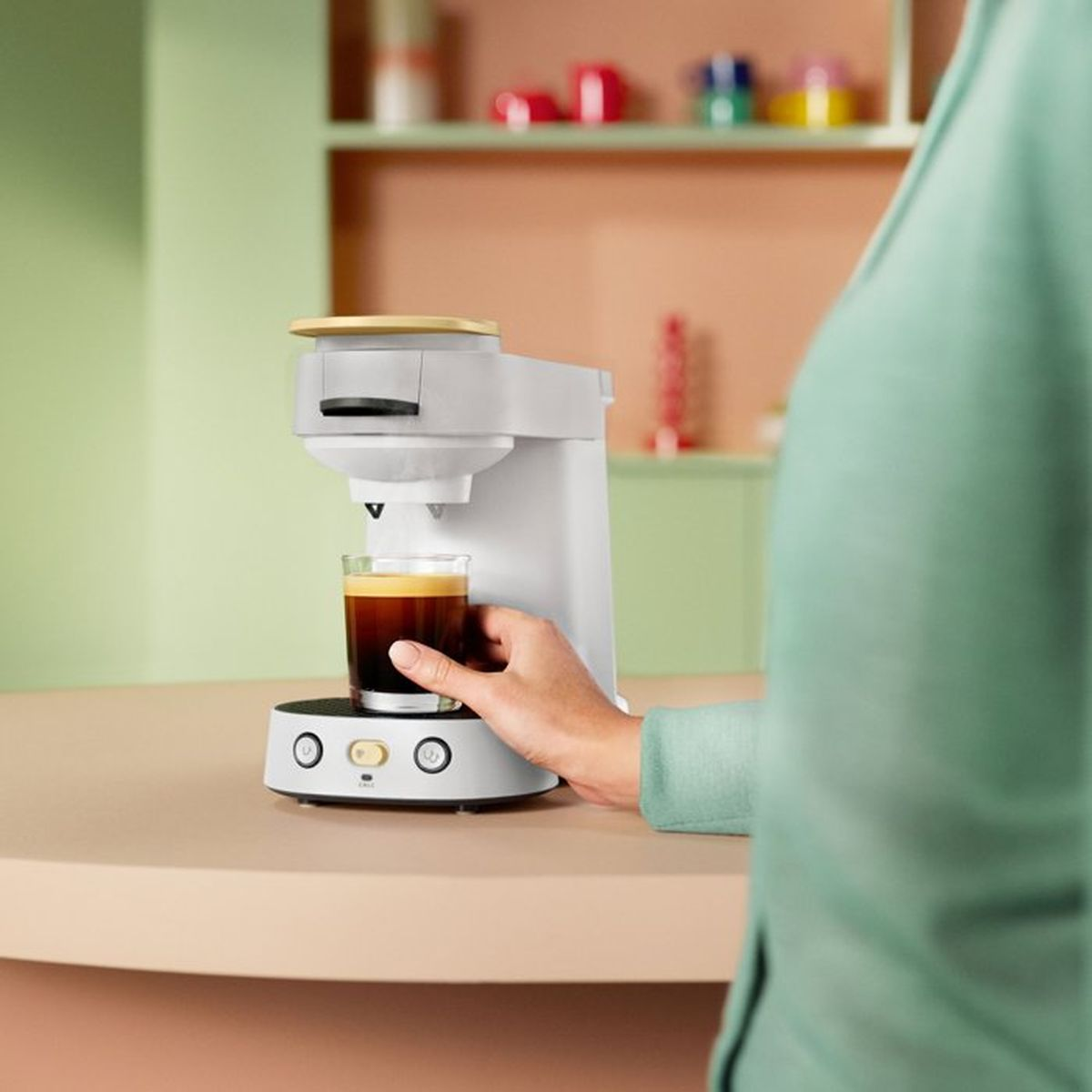 Person giesst Kaffee aus einer weissen Kaffeemaschine in ein Glas auf einer Theke.