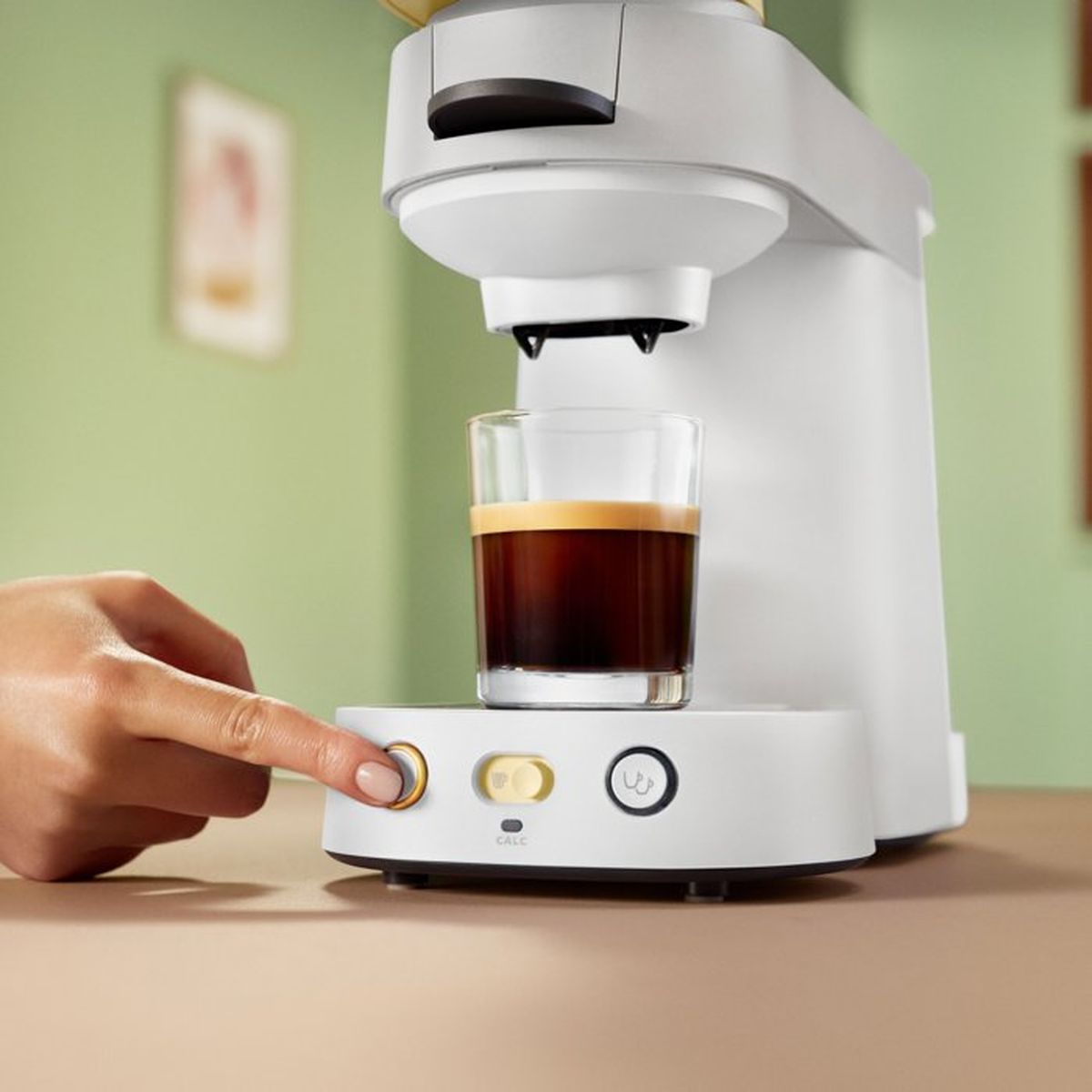 Eine Hand drückt den Knopf einer weißen Kaffeemaschine, auf der eine Tasse frischer Kaffee steht.