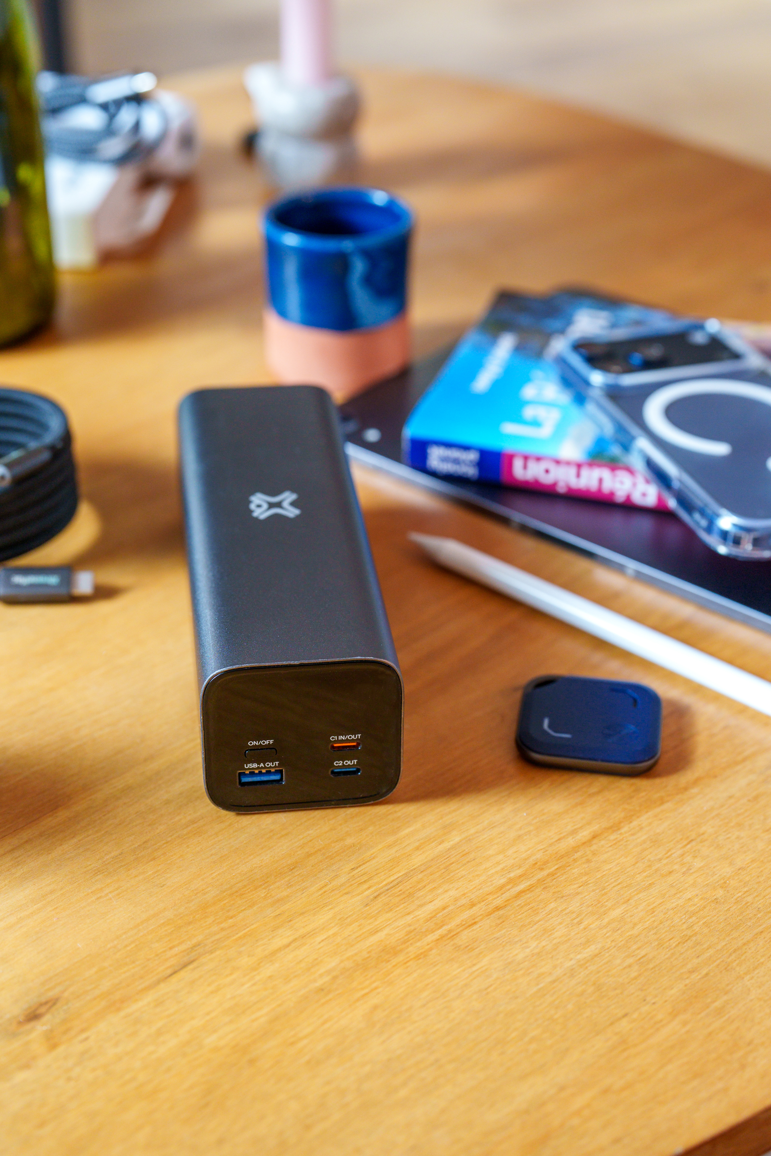 Eine schwarze Powerbank auf einem Holztisch, umgeben von einem Stift, einer blauen Tasse und einem Smartphone.