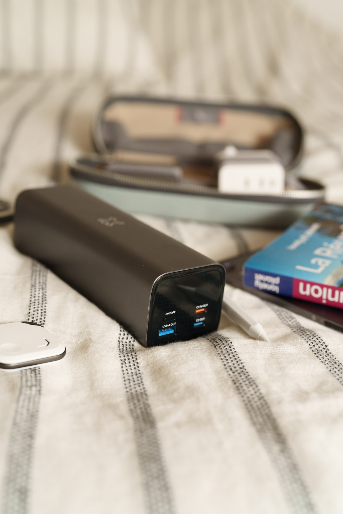 Eine schwarze Powerbank mit USB-Anschlüssen liegt auf einem Bett neben einem Buch und einer grauen Tasche.