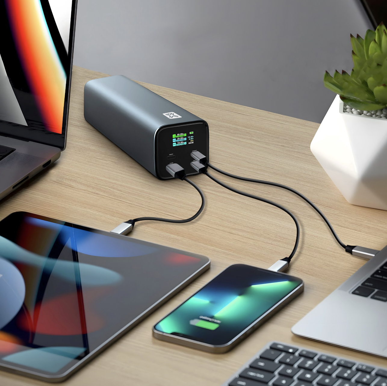 Eine graue Powerbank auf einemHolztisch lädt über Kabel ein Tablet, ein Smartphone und einen Laptop.