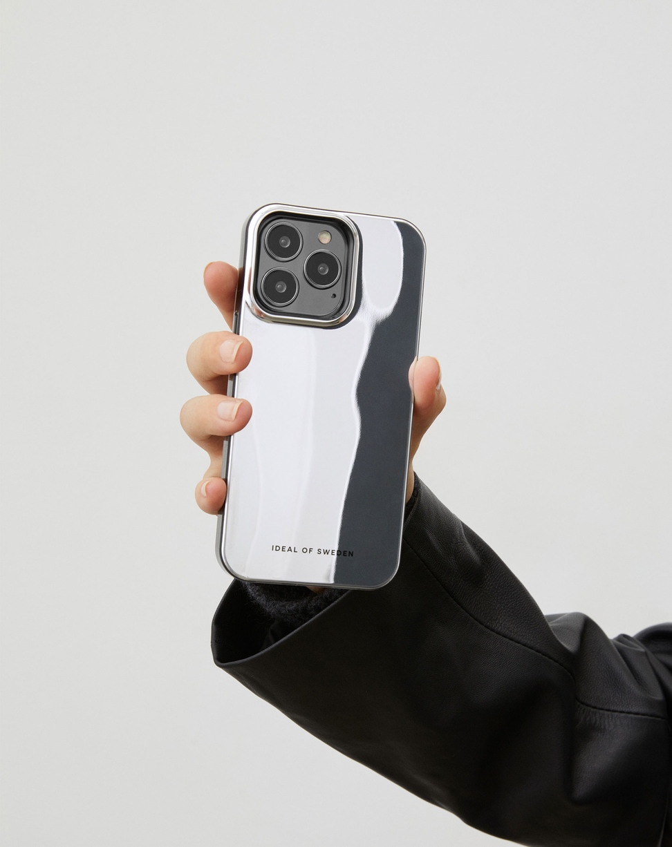 Eine Hand in einer schwarzen Jacke hält ein Smartphone mit spiegelnder, silberner Hülle vor hellem Hintergrund.