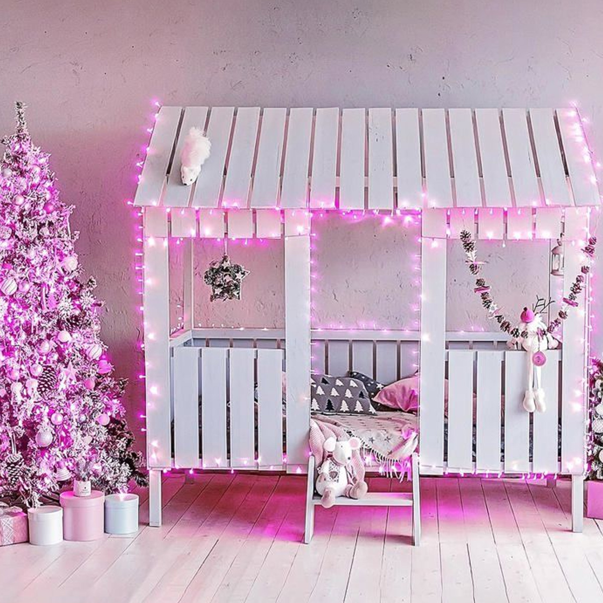Ein weißes Hausbett mit rosa Lichterketten und ein pinker Weihnachtsbaum im Raum.