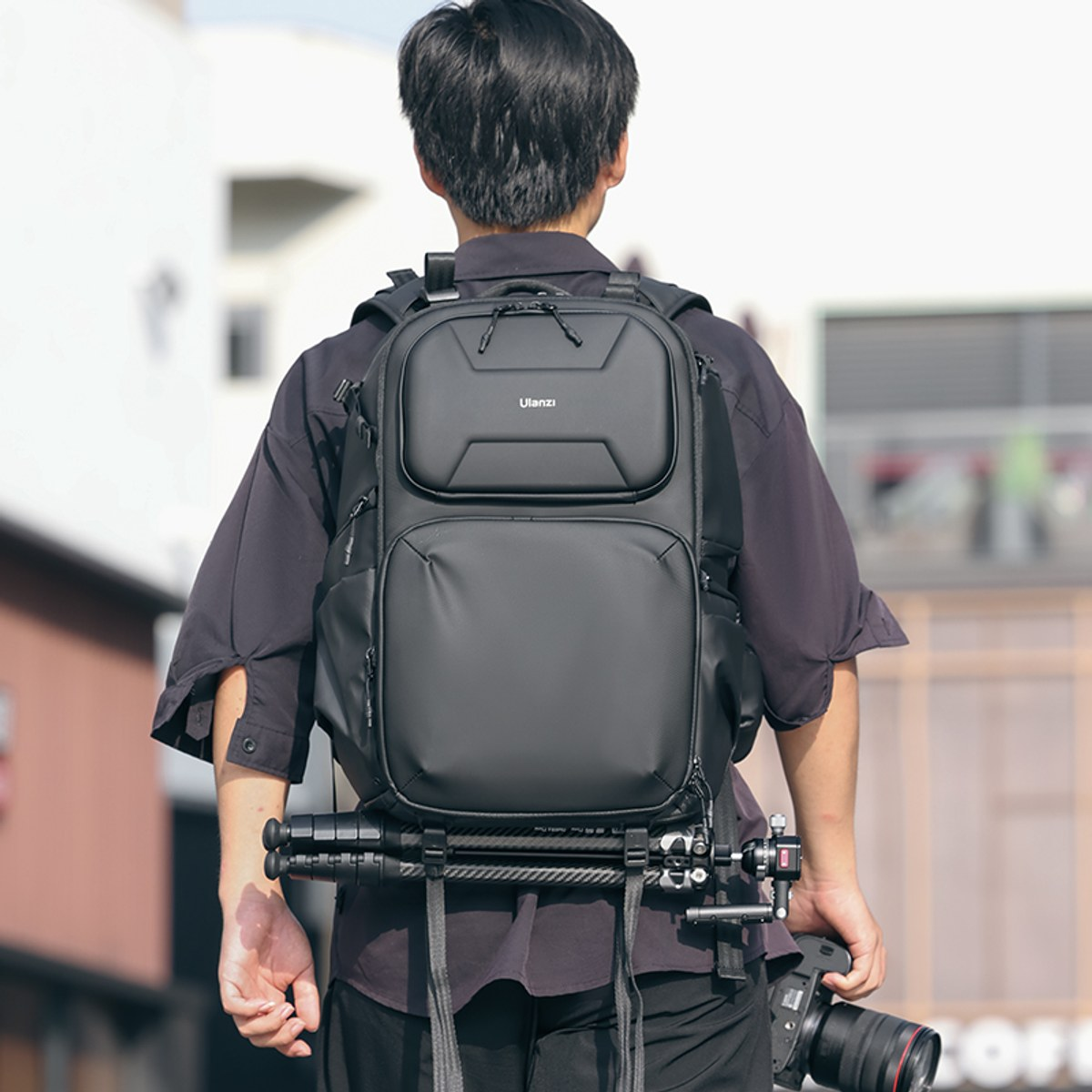 Siyah gömlekli ve altında tripod olan büyük siyah Ulanzi sırt çantalı bir adam kamera tutuyor.