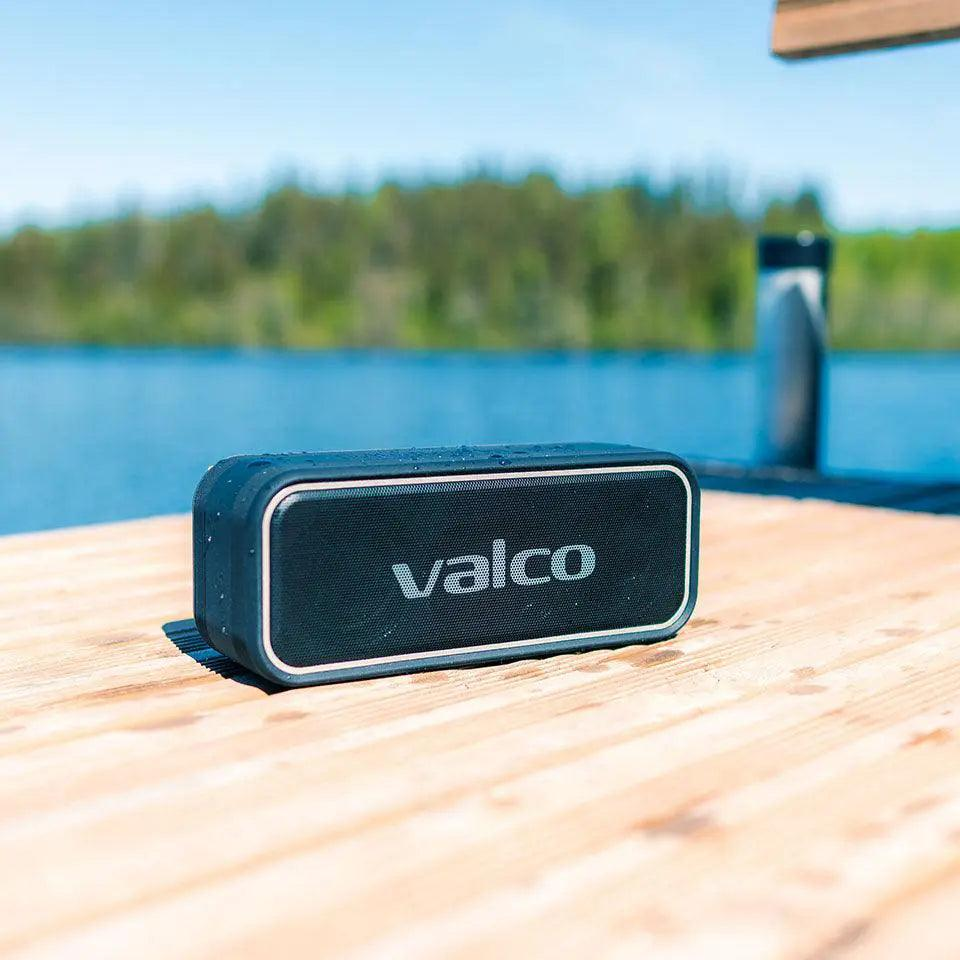 Ein schwarzer Valco Bluetooth-Lautsprecher auf einem Holzsteg vor einem blauen See und grüne Bäume im Hintergrund.
