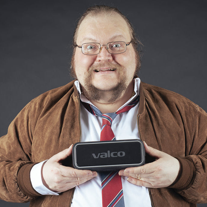 Ein Mann mit Brille und Bart hält lächelnd einen kleinen schwarzen Valco-Lautsprecher in die Kamera.