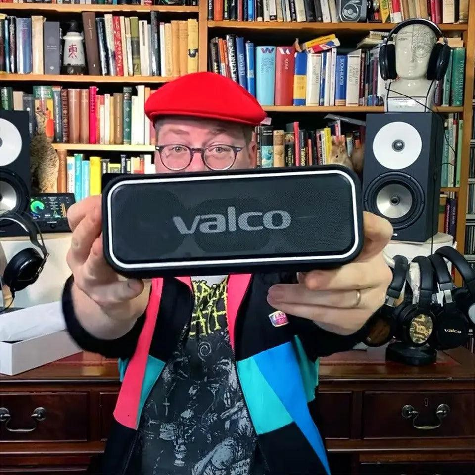 Ein Mann mit roter Mütze und Brille hält einen Valco Lautsprecher vor eine mit Büchern gefüllte Wand.