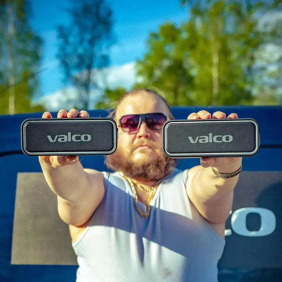 Ein bärtiger Mann mit Sonnenbrille hält zwei Valco Lautsprecher vor einem blauen Auto und grünen Bäumen in die Höhe.