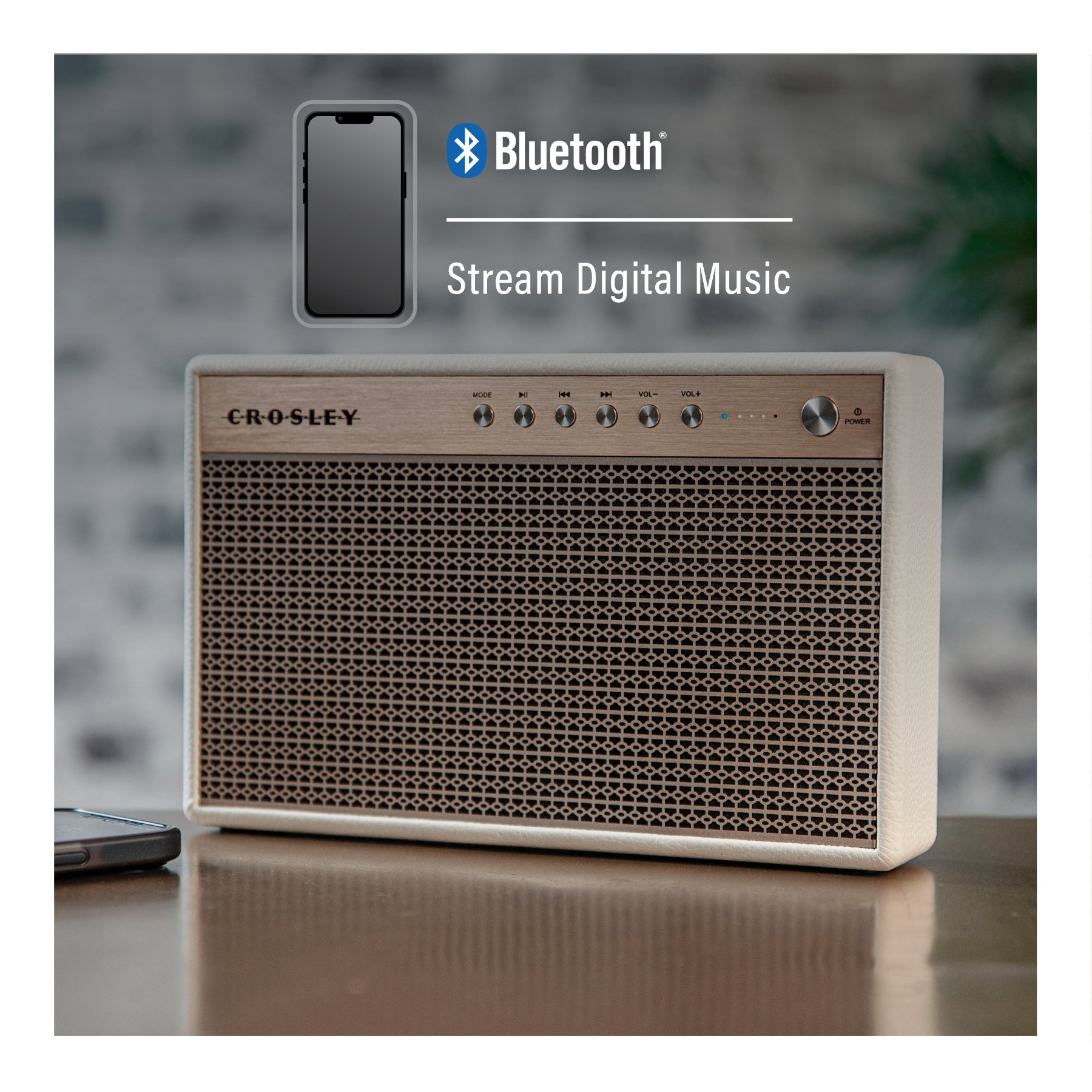 Ein cremefarbener Crosley Bluetooth-Lautsprecher steht auf einem Holztisch vor einem verschwommenen Hintergrund.