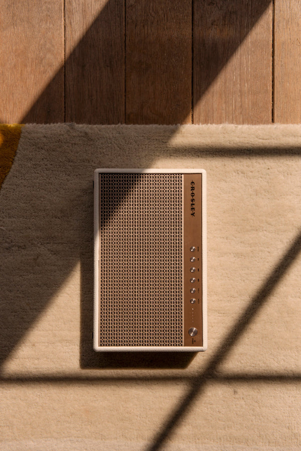 Ein cremefarbener Crosley-Lautsprecher liegt auf einem Teppich, im harten Sonnenlicht mit kantigen Schatten.