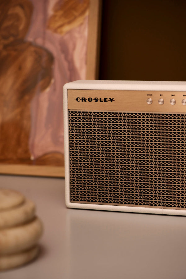 Ein weißer Crosley-Lautsprecher mit goldenem Gitter steht auf einem Tisch vor einem abstrakten Gemälde.