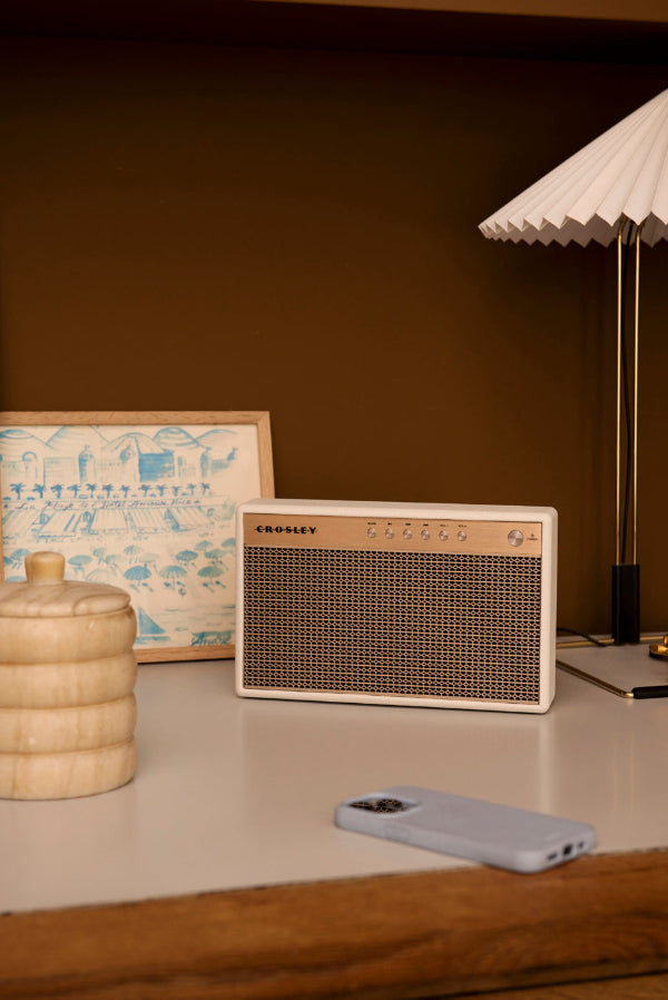 Ein weißer Crosley-Lautsprecher, eine geriffelte Lampe, ein Bild, ein Behälter und ein Smartphone auf einem Tisch.