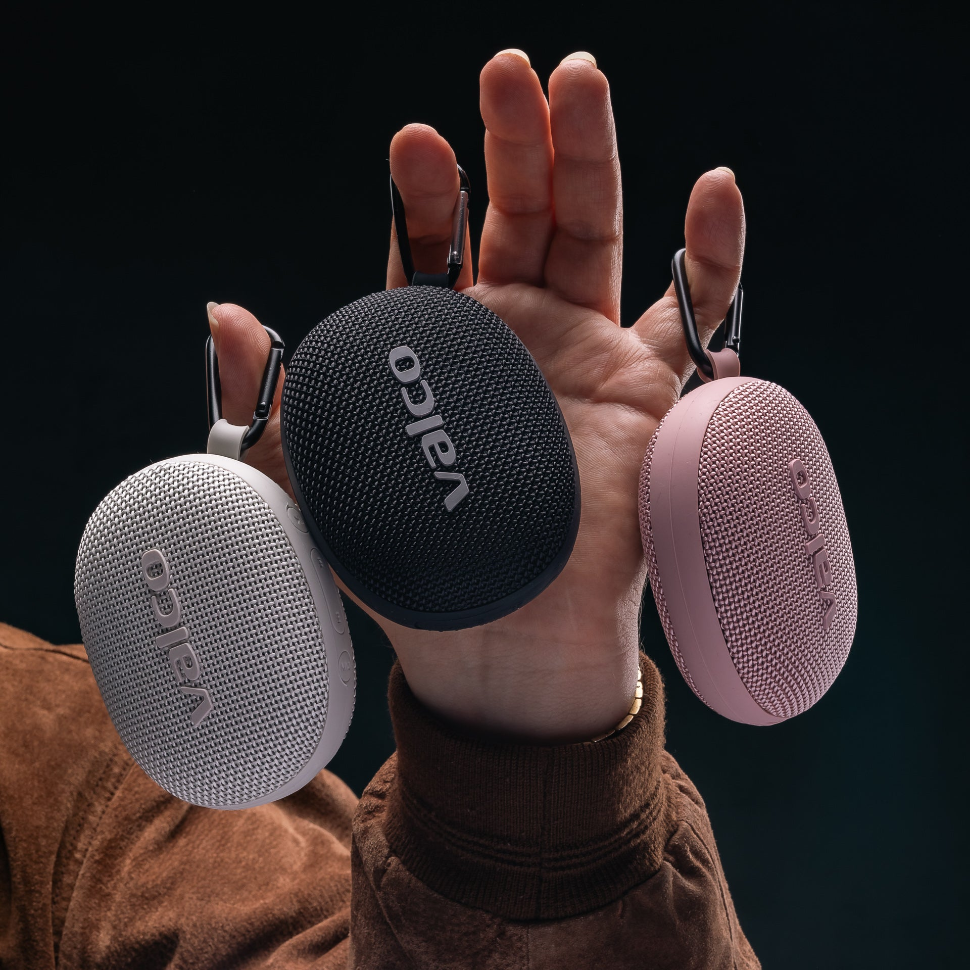 Eine Hand hält drei ovale Valco-Lautsprecher in den Farben Weiß, Schwarz und Rosa mit Karabinerhaken.