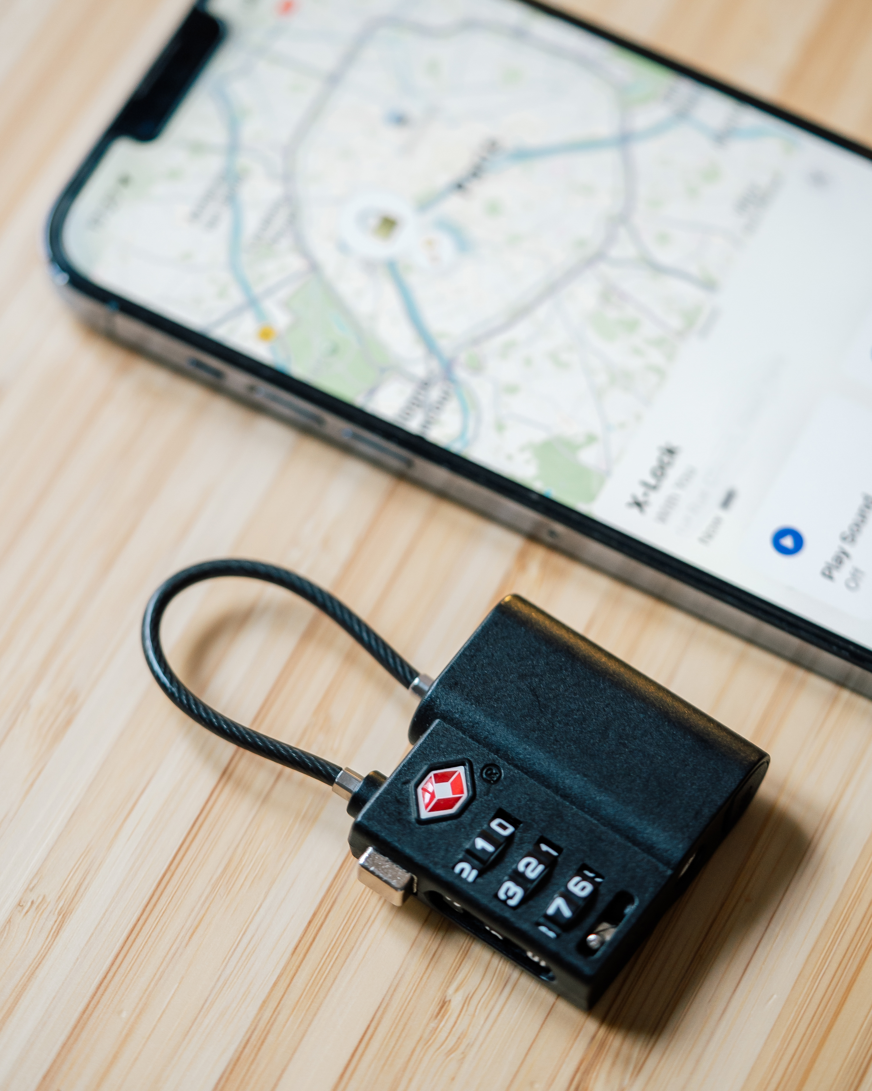Ein schwarzes Kofferschloss mit Zahlencode liegt auf einem Holztisch neben einem Smartphone mit Kartennavigation.