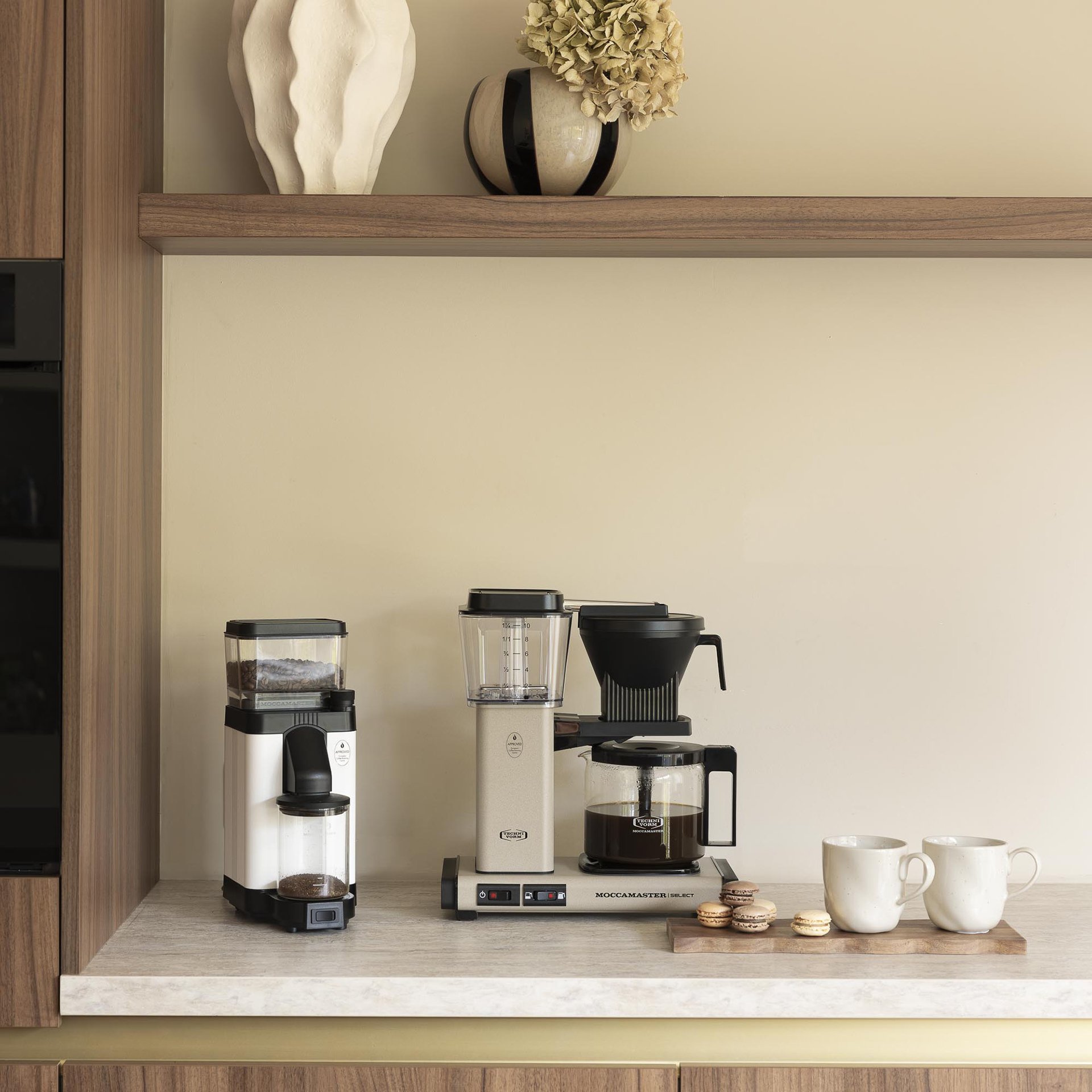 Comptoir de cuisine beige avec cafetière, moulin, tasses et macarons, sous une étagère en bois avec des vases.