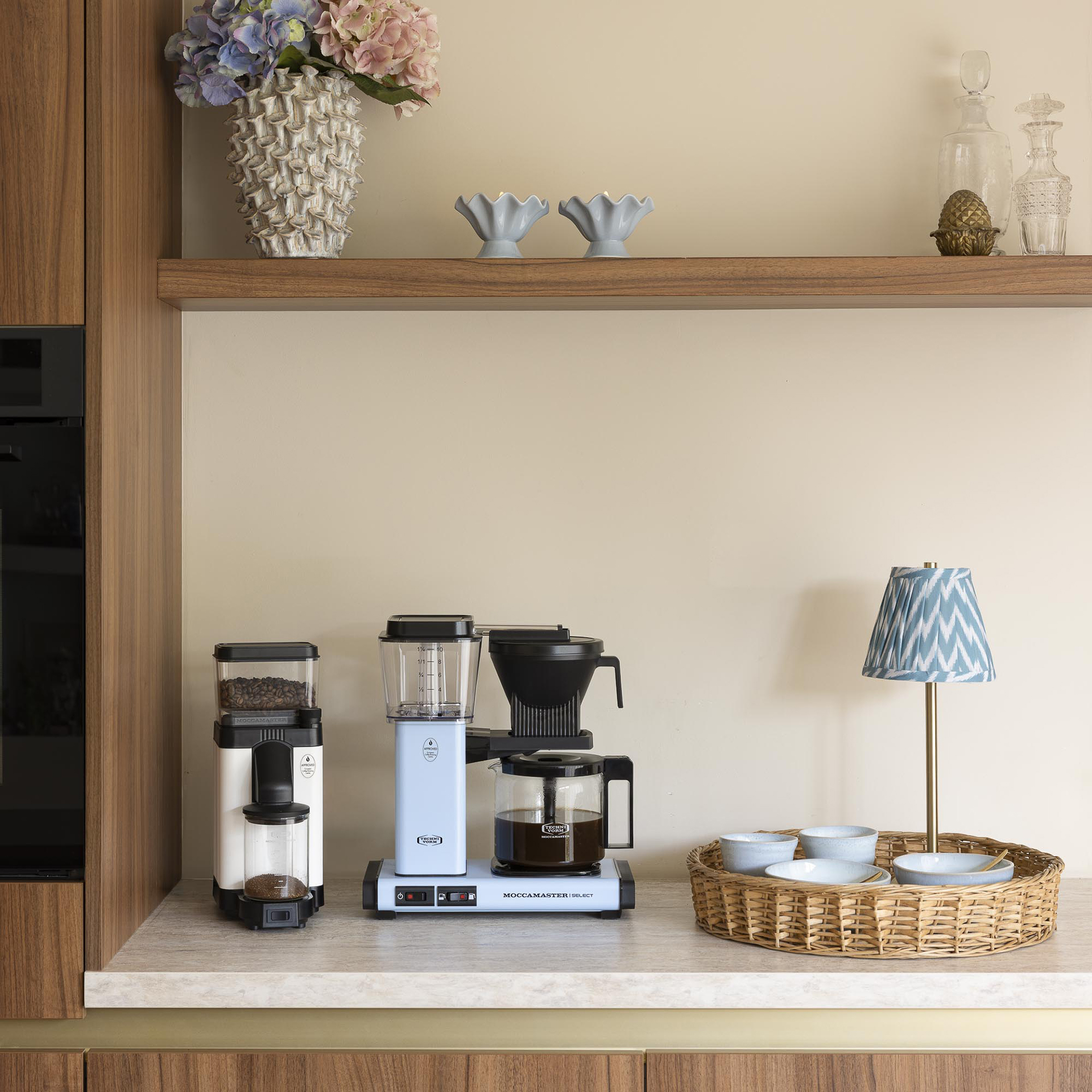 Cafetière et moulin bleu clair sur un comptoir avec un plateau en osier, des bols et une petite lampe.