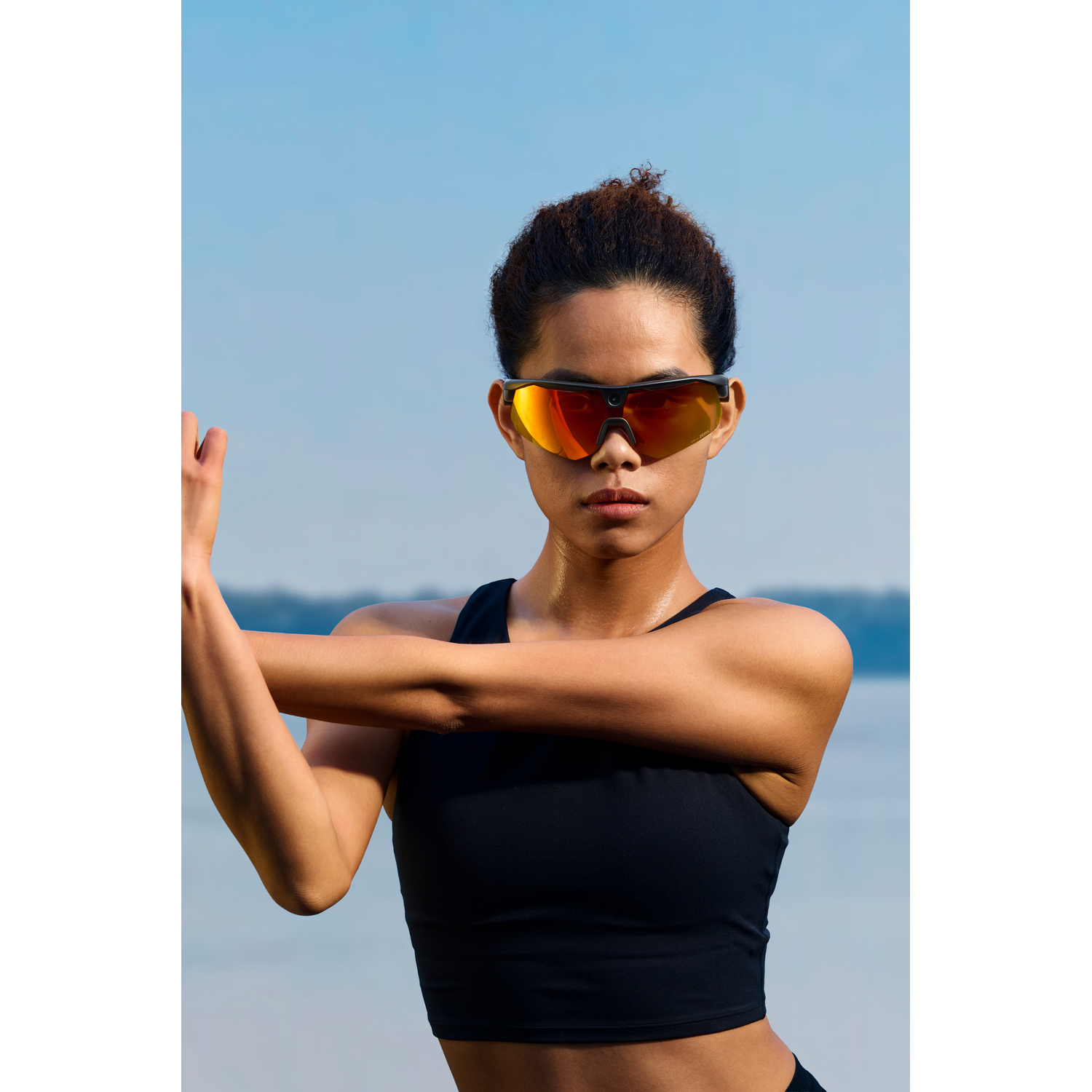 Eine Frau in Sportkleidung und mit orangeroten Sonnenbrillen dehnt sich draußen vor klarem Himmel und Wasser.