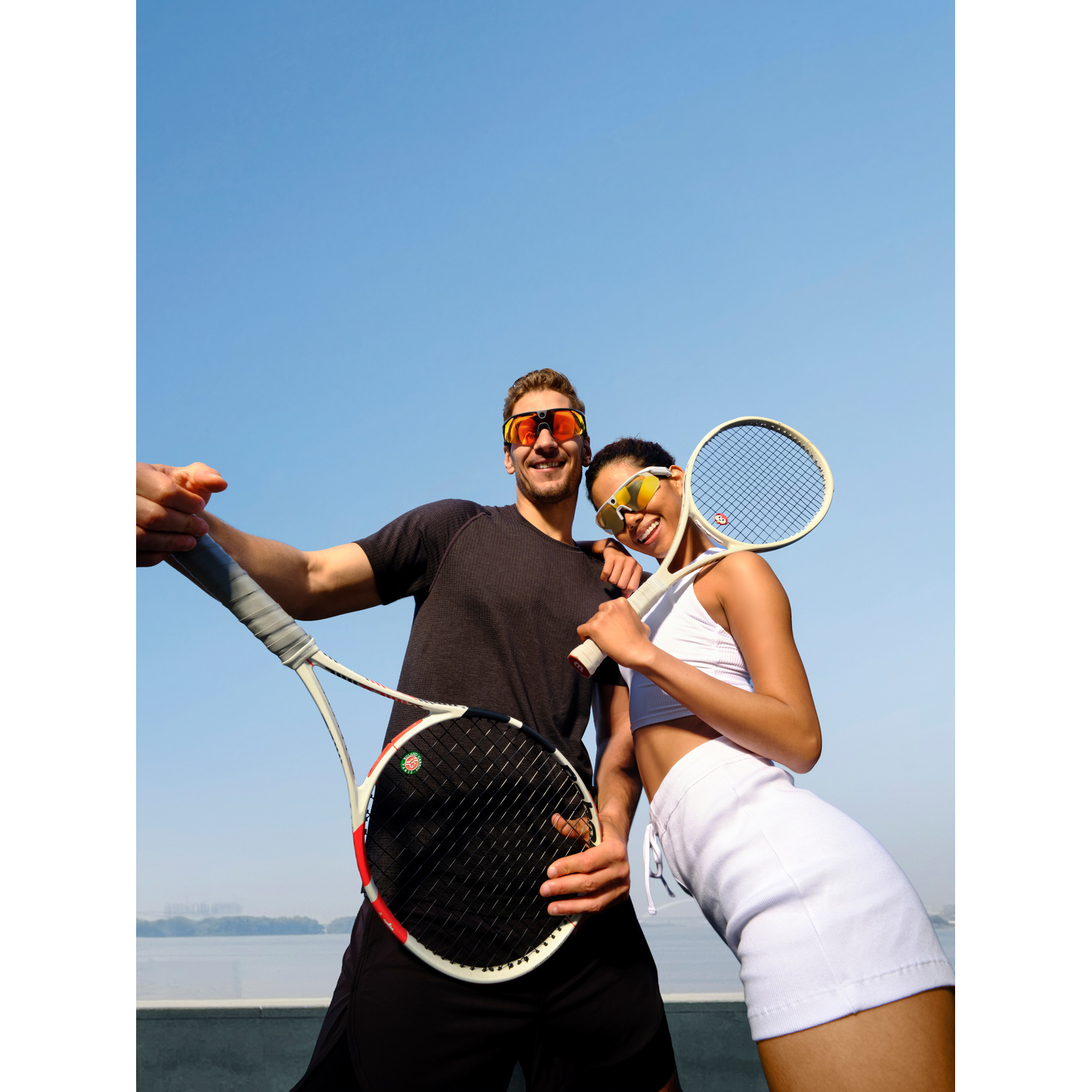 Ein Mann und eine Frau in Sportkleidung halten Tennisschläger und lächeln vor blauem Himmel.