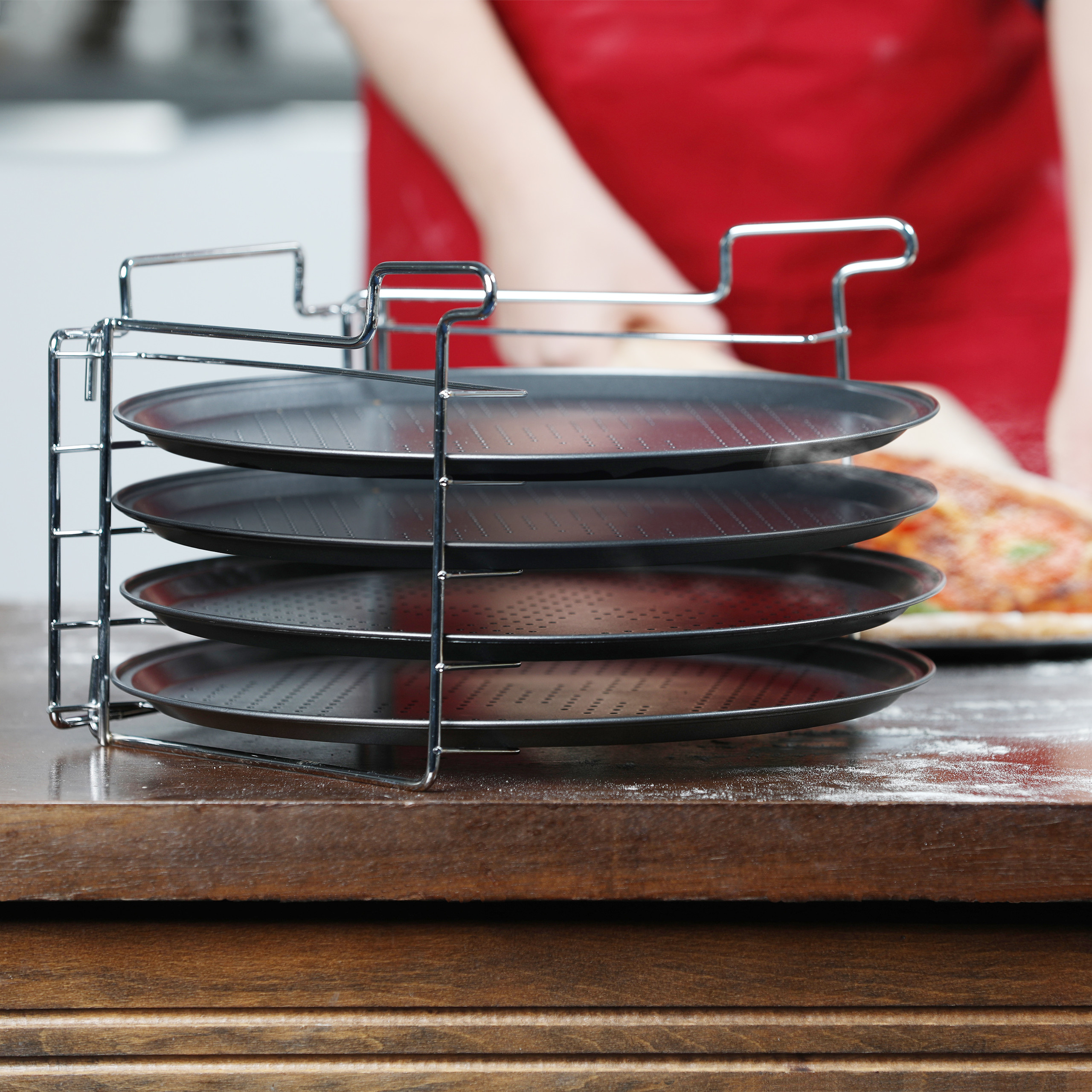 Ein Metallgestell hält vier schwarze Pizzableche auf einem Holztisch, im Hintergrund eine Person in roter Schürze.