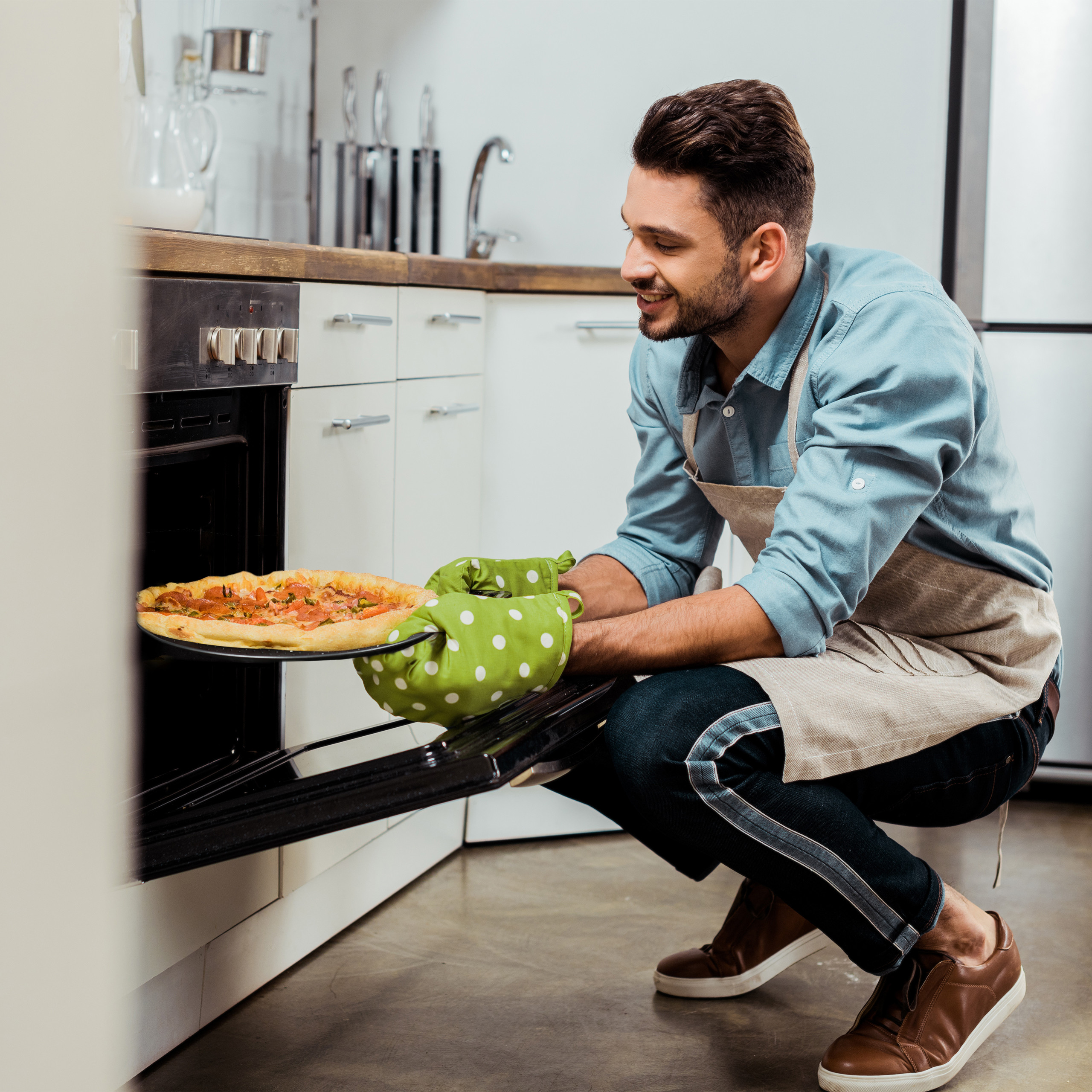 Ein lächelnder Mann in Schürze und Ofenhandschuhen nimmt eine frisch gebackene Pizza aus dem Ofen in der Küche.