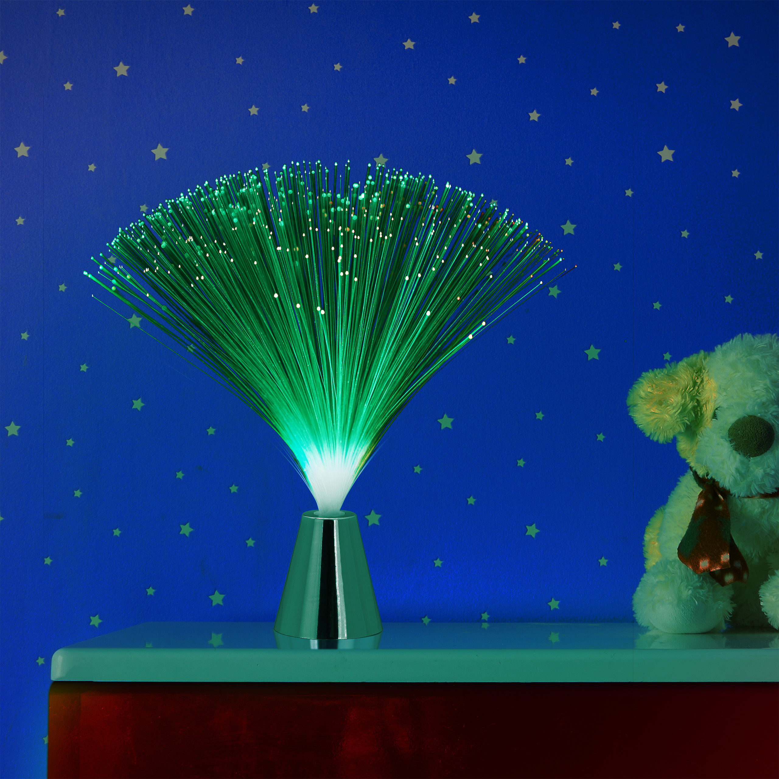 Eine leuchtende grüne Faseroptiklampe und ein Plüschhund stehen auf einem Regal vor einer blauen Wand mit Sternen.