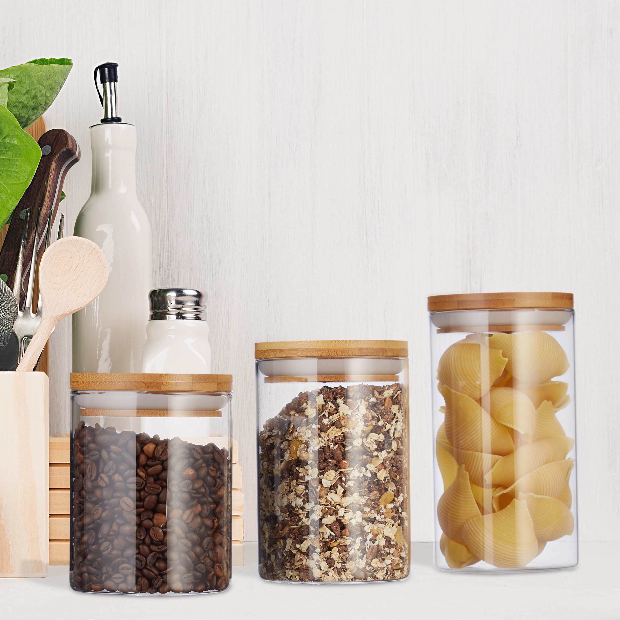 Glasbehälter mit Kaffeebohnen, Müsli und Nudeln, daneben Küchenutensilien auf weißem Grund.