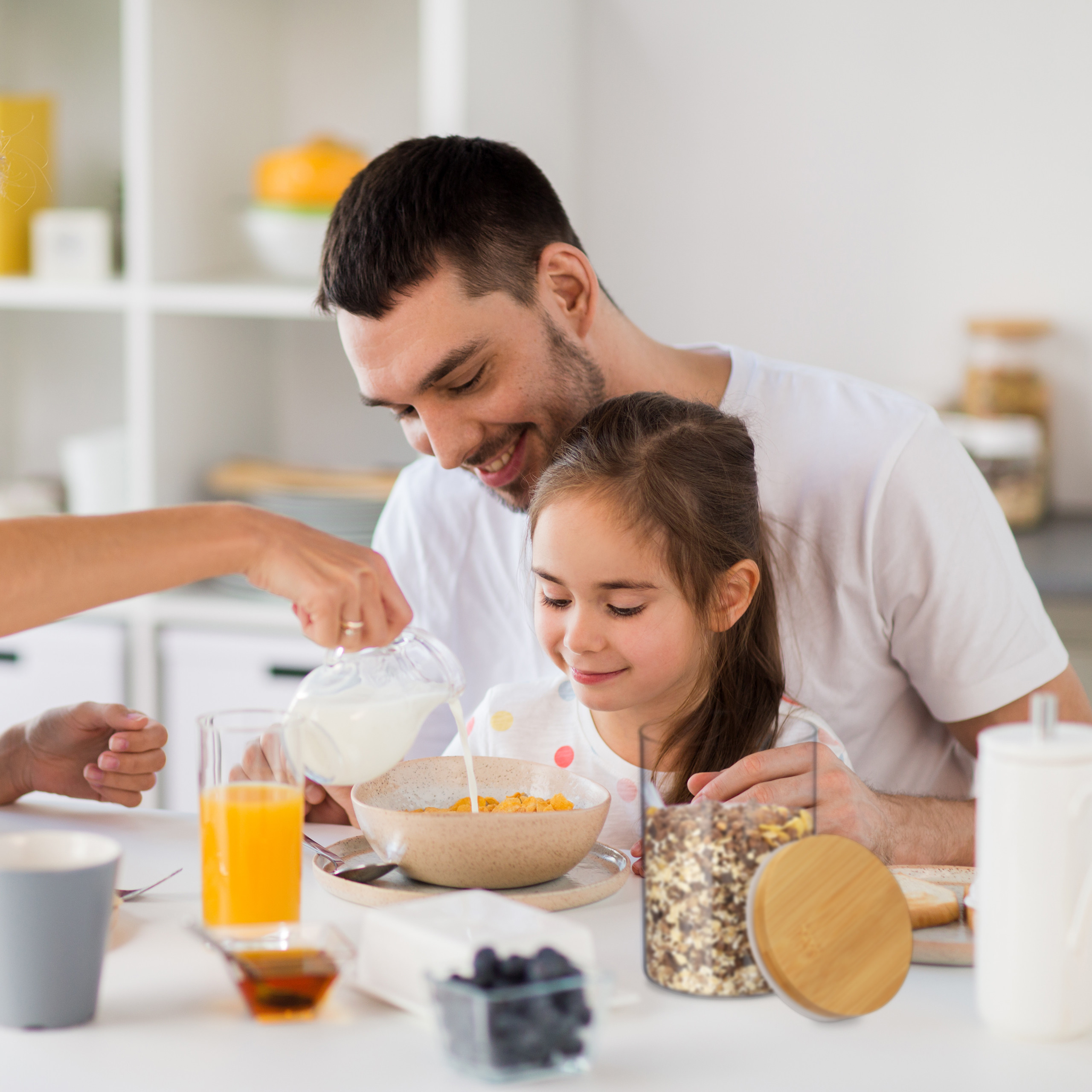 Ein Vater lächelt, während jemand Milch in die Müslischüssel seiner Tochter gießt.