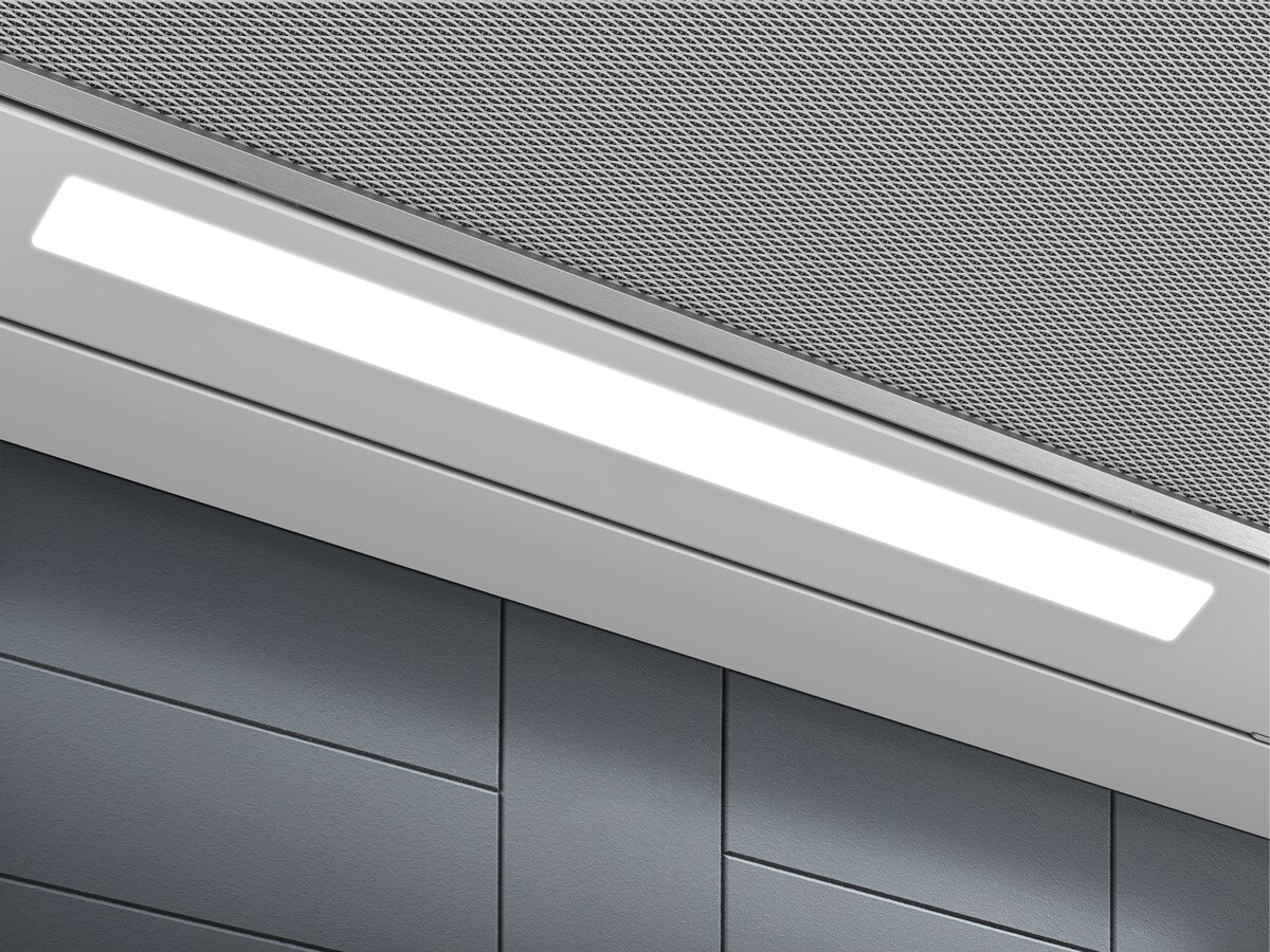 Parte inferior de una campana extractora con tira LED brillante y rejilla metálica sobre pared gris.