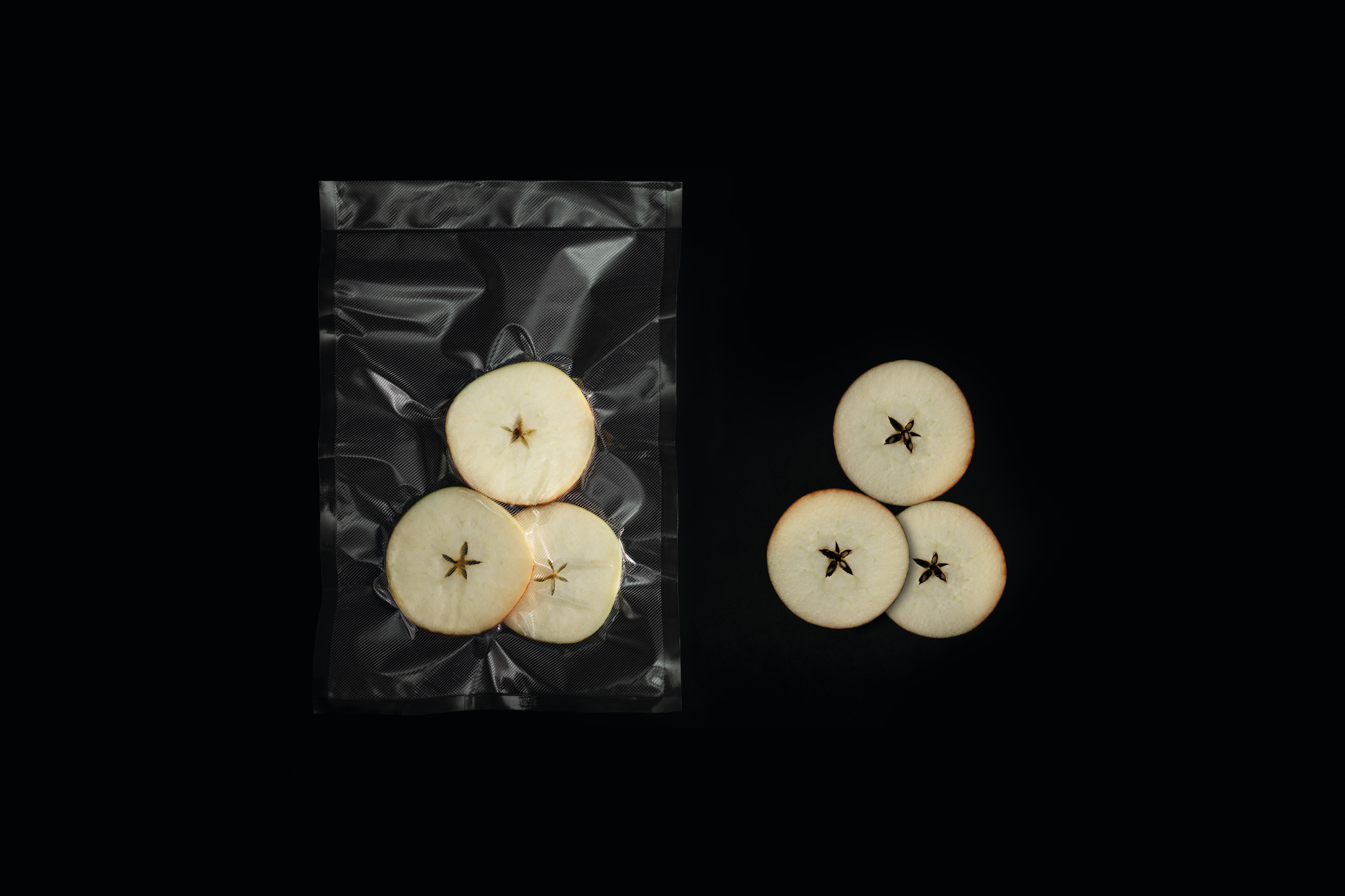 Tres rodajas de manzana en una bolsa al vacío junto a tres rodajas sueltas sobre fondo negro.