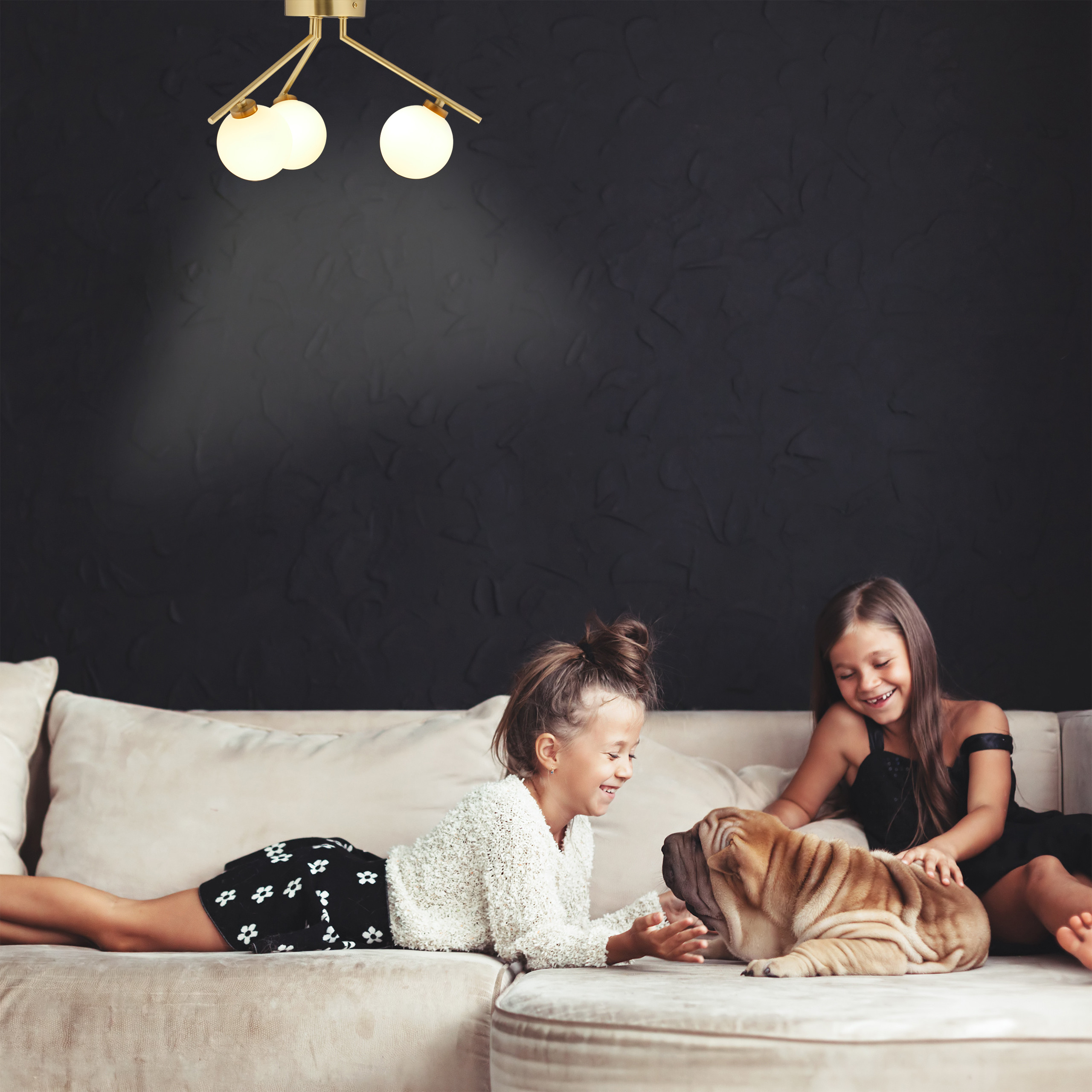 Zwei Mädchen spielen auf einer beigen Couch mit einem Shar-Pei-Welpen unter einer goldenen Deckenlampe vor einer dunklen Wand.