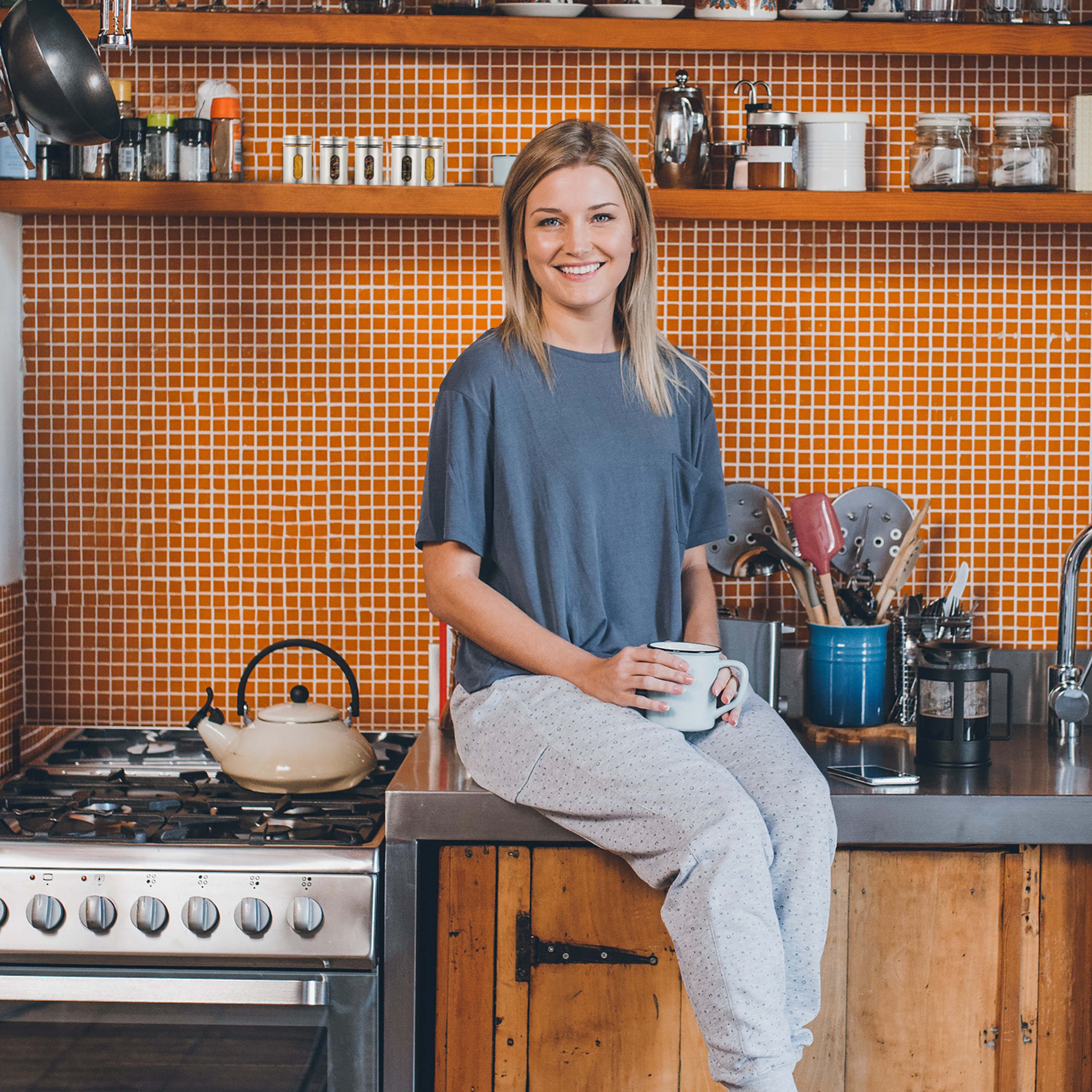 Eine blonde Frau sitzt lächelnd mit einer Tasse in einer K üche vor einer orangefarbenen Wand.