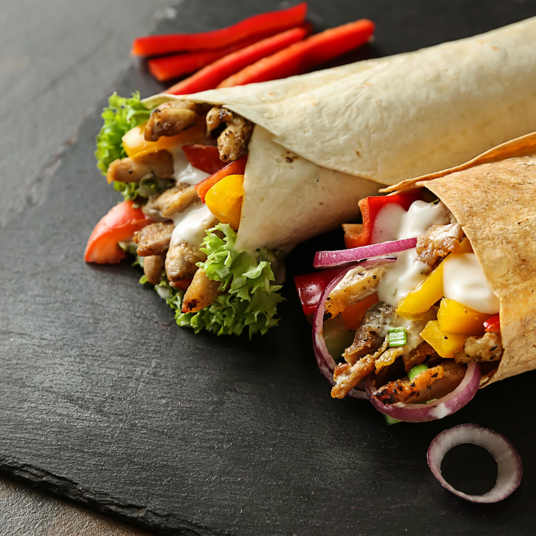 Zwei Hähnchenwraps mit Salat, Paprika, roten Zwiebeln und Sauce auf schwarzem Schiefer.