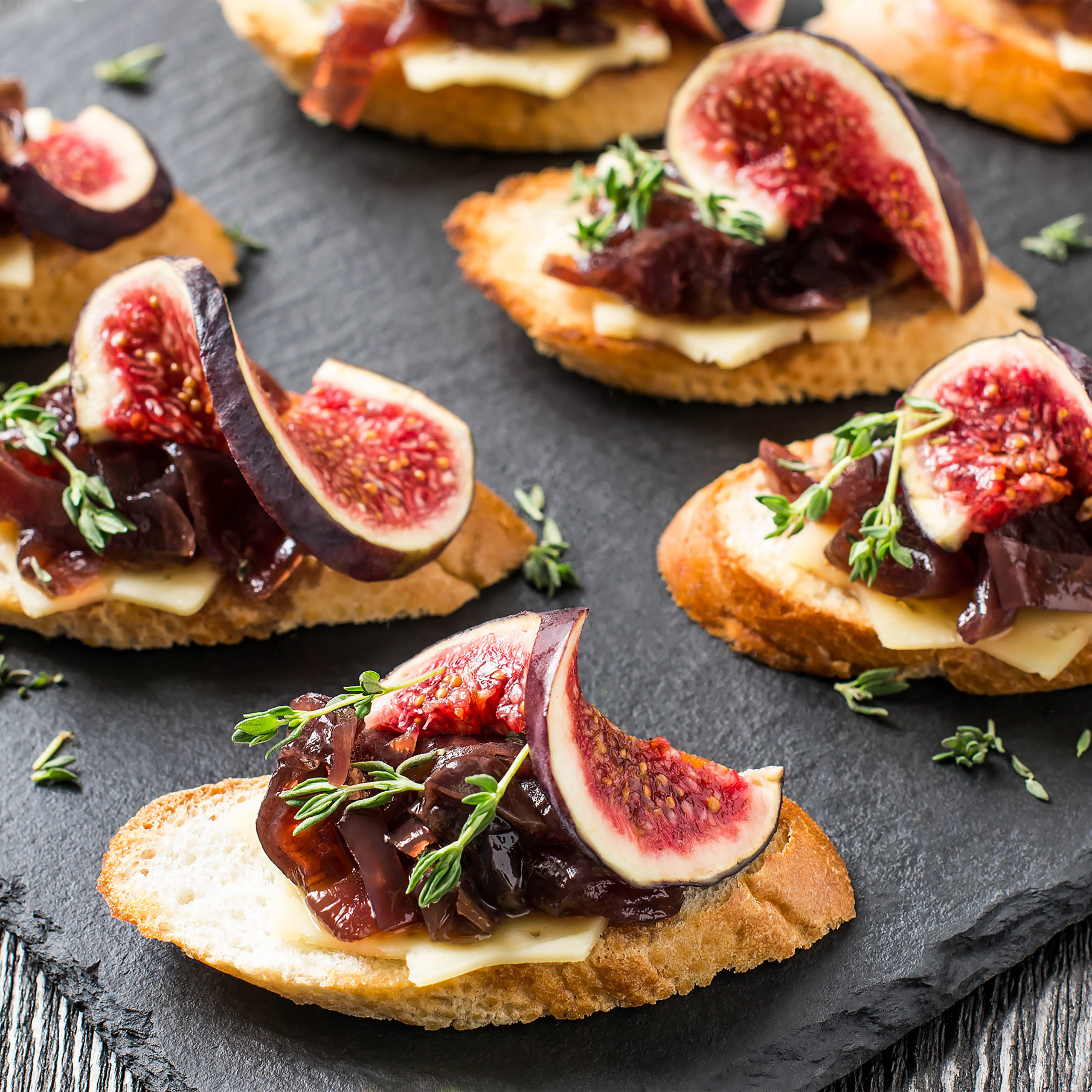 Knusprige Bruschetta mit geschmolzenem Käse, Feigen, Zwiebeln und Thymian auf einem schwarzen Brett.