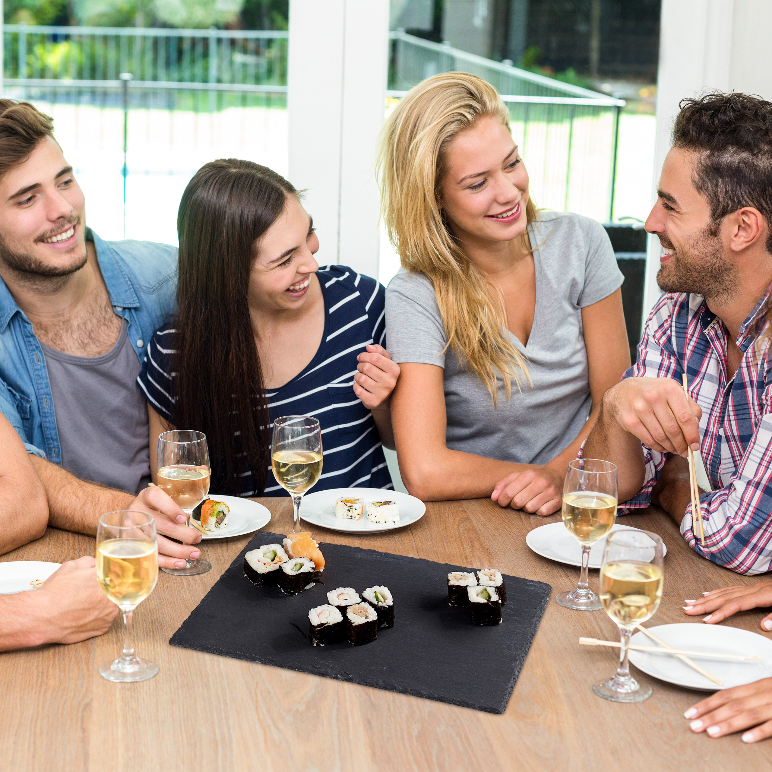 Vier Freunde lachen und essen Sushi mit Weißwein an einem Holztisch drinnen.