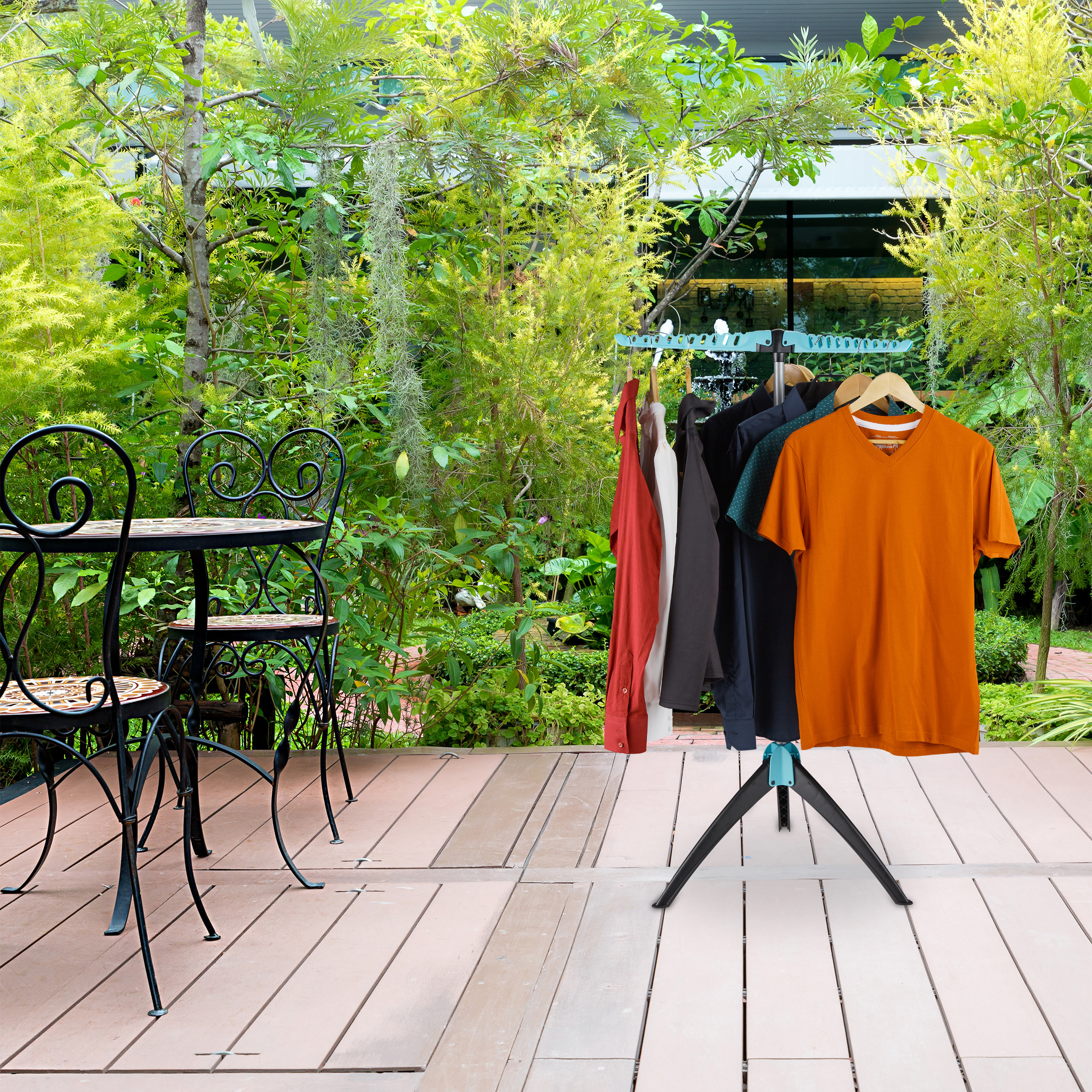 Wäscheständer mit Kleidung auf einer Holzterrasse vor grünem Garten mit Esstisch und Stühlen.