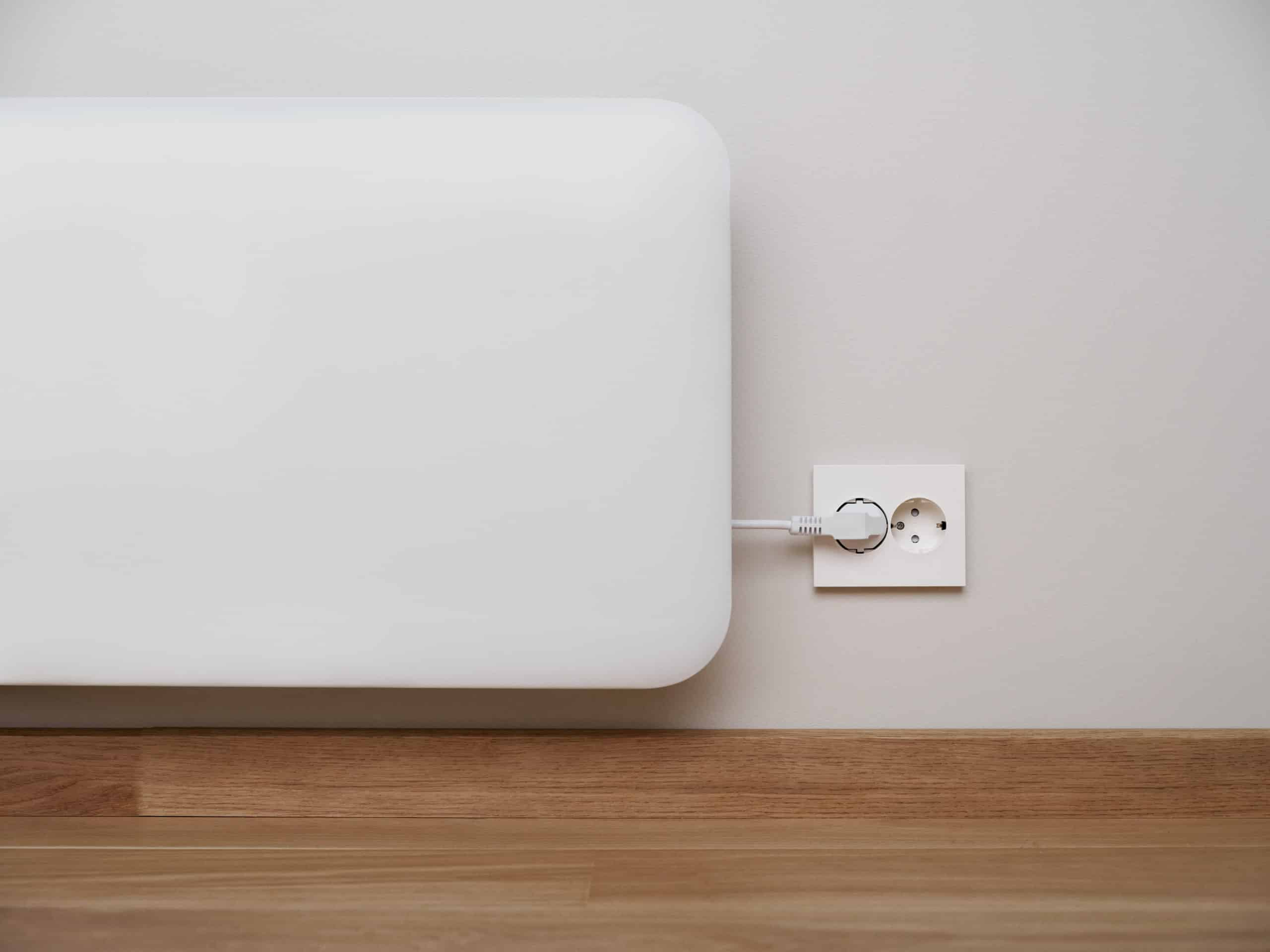 Un riscaldatore elettrico bianco su una parete grigia, collegato a una presa di corrente sopra un pavimento in legno.