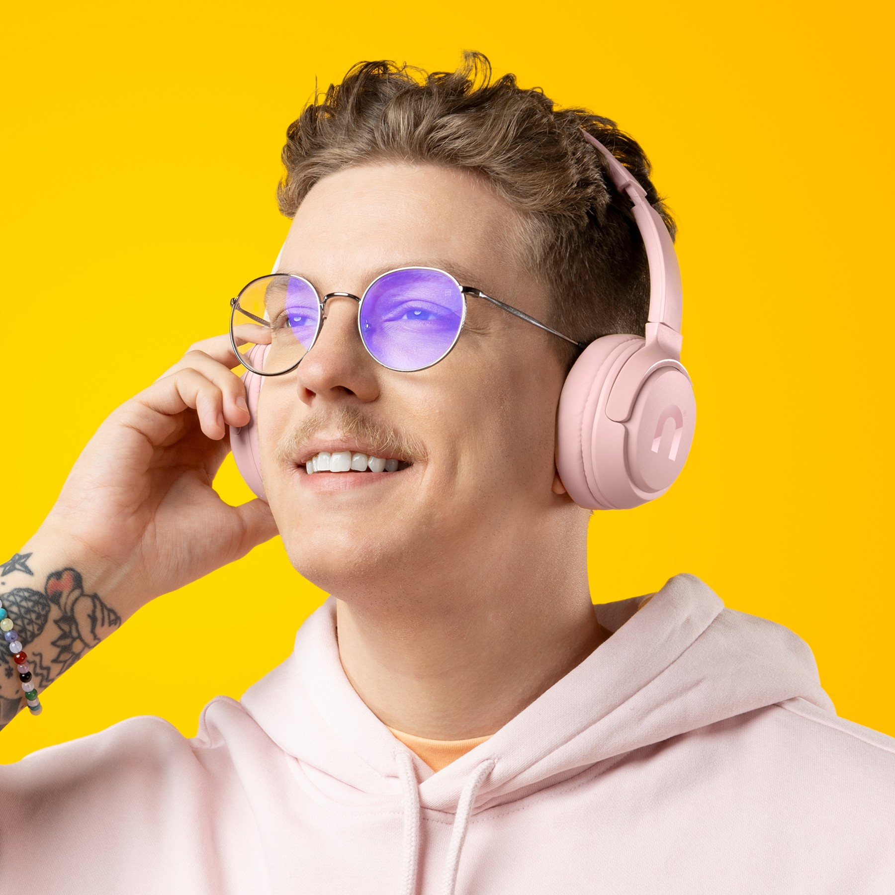 Ein Mann mit rosa Kopfhörern, runden Brillen und einem rosa Hoodie lächelt vor einem gelben Hintergrund.