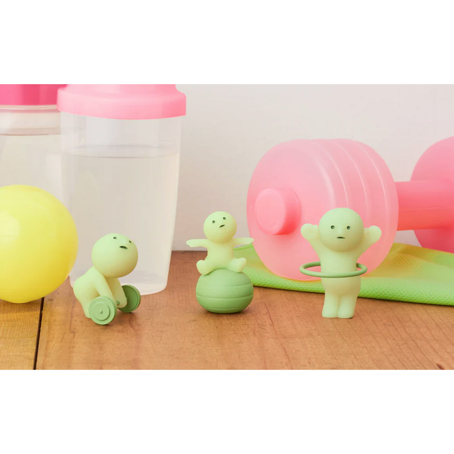 Drei kleine grüne Figuren trainieren mit rosa Hanteln, einem Ball und einem Hula-Hoop-Reifen auf Holz.