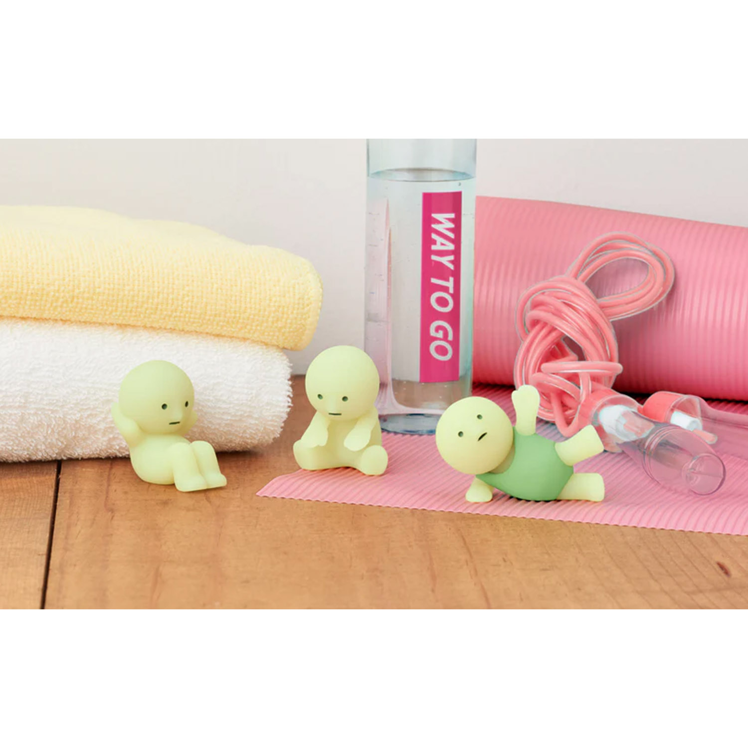 Drei kleine grüne Figuren üben neben Handtüchern, einer Wasserflasche, einer Yogamatte und einem Seil auf einem Holztisch.