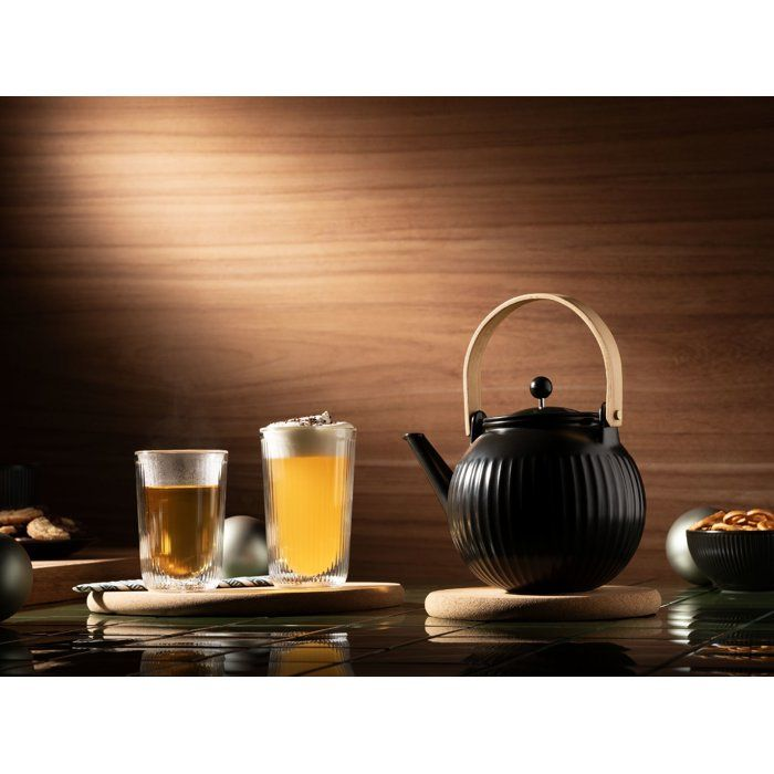 Una tetera negra y dos vasos de té en posavasos de madera contra una pared de madera.
