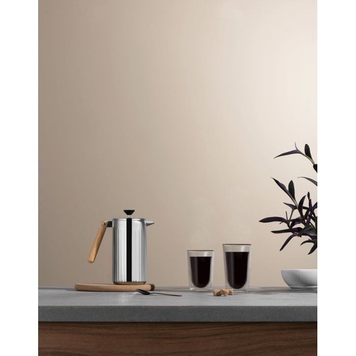 Eine silberne French Press und zwei Gläser Kaffee stehen auf einer grauen Arbeitsplatte neben einer kleinen Pflanze.