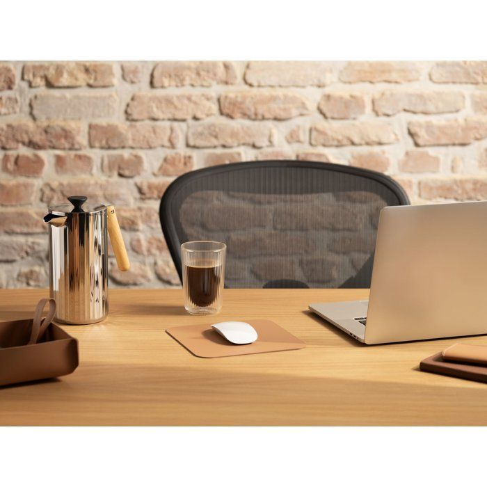 Ein Holztisch mit Laptop, Kaffeebereiter, Glas Kaffee und Mauspad vor einer Ziegelwand.