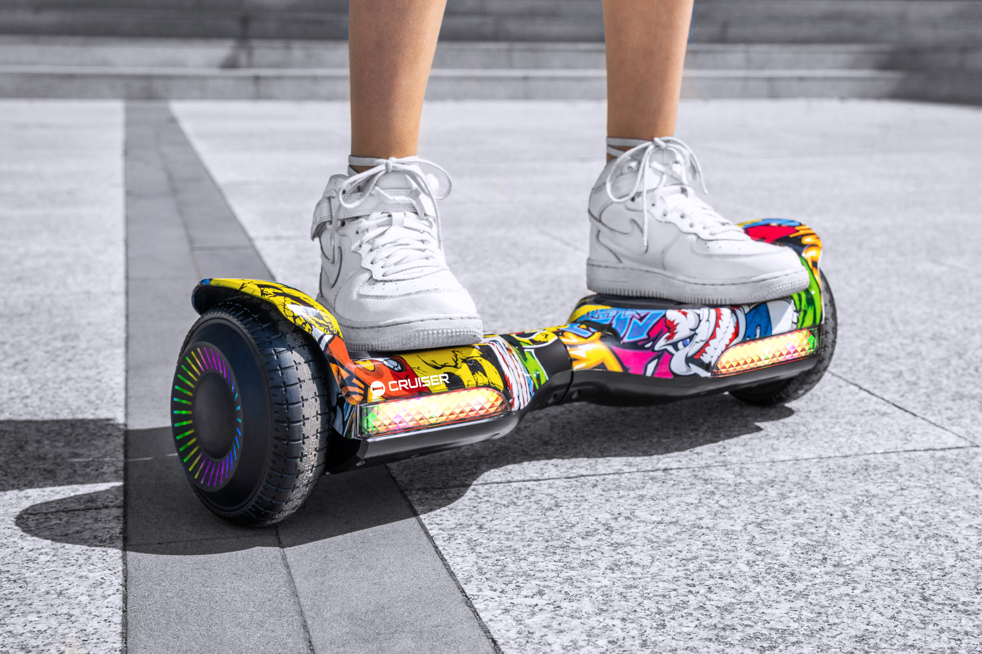Osoba w białych tenisówkach stoi na kolorowej desce typu hoverboard ze świecącymi światłami na szarym chodniku.