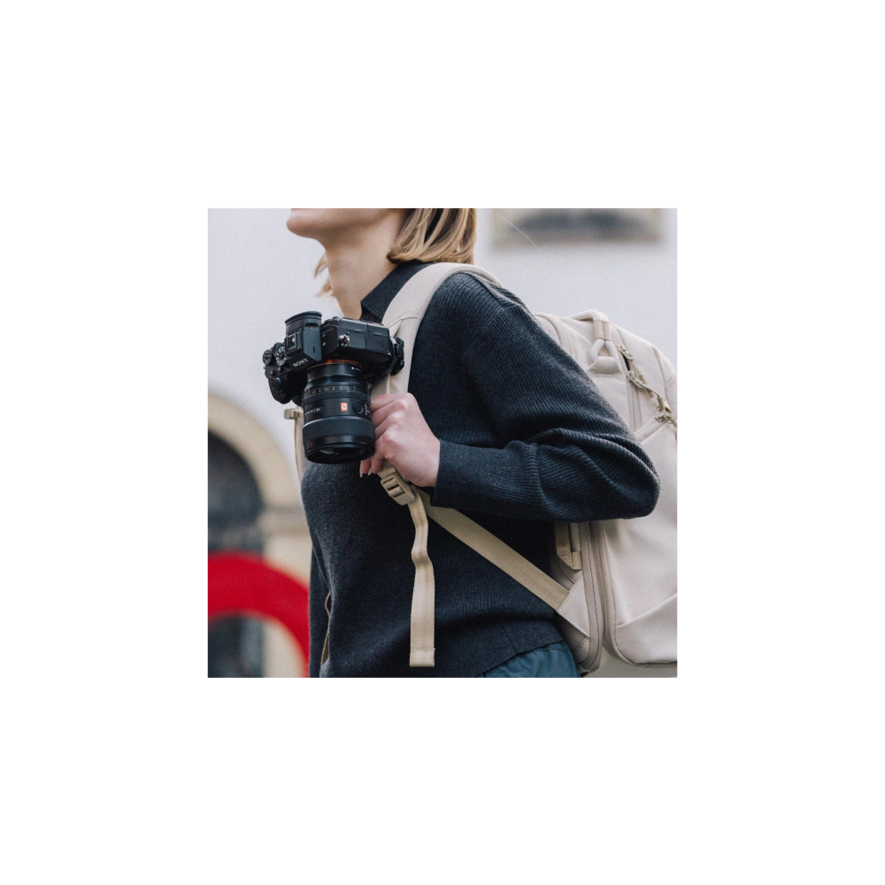 Eine Frau im grauen Pullover mit beige Rucksack hält eine Kamera in der Hand.