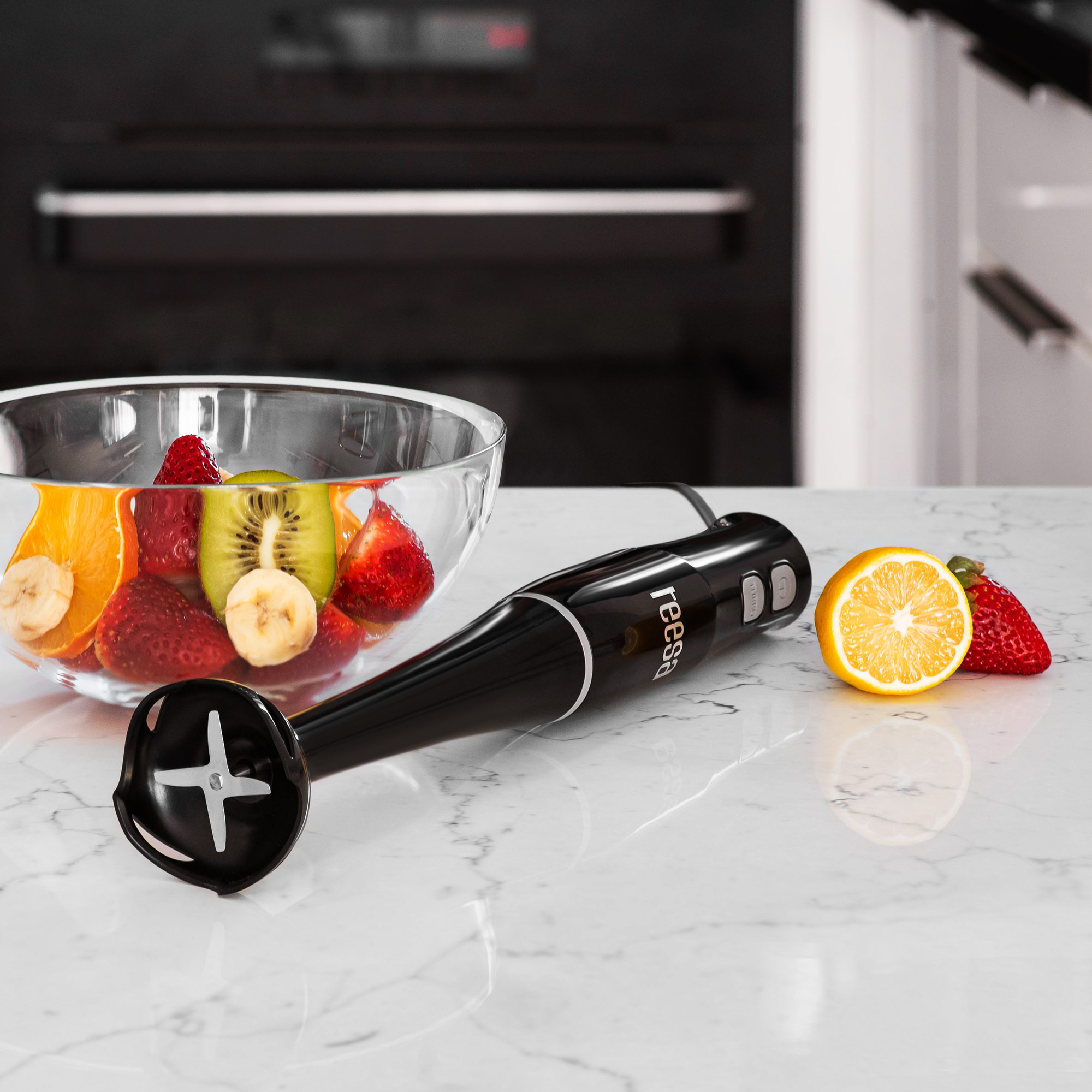 Czarny blender ręczny leży na marmurowym blacie obok miski z owocami i połówki cytryny.