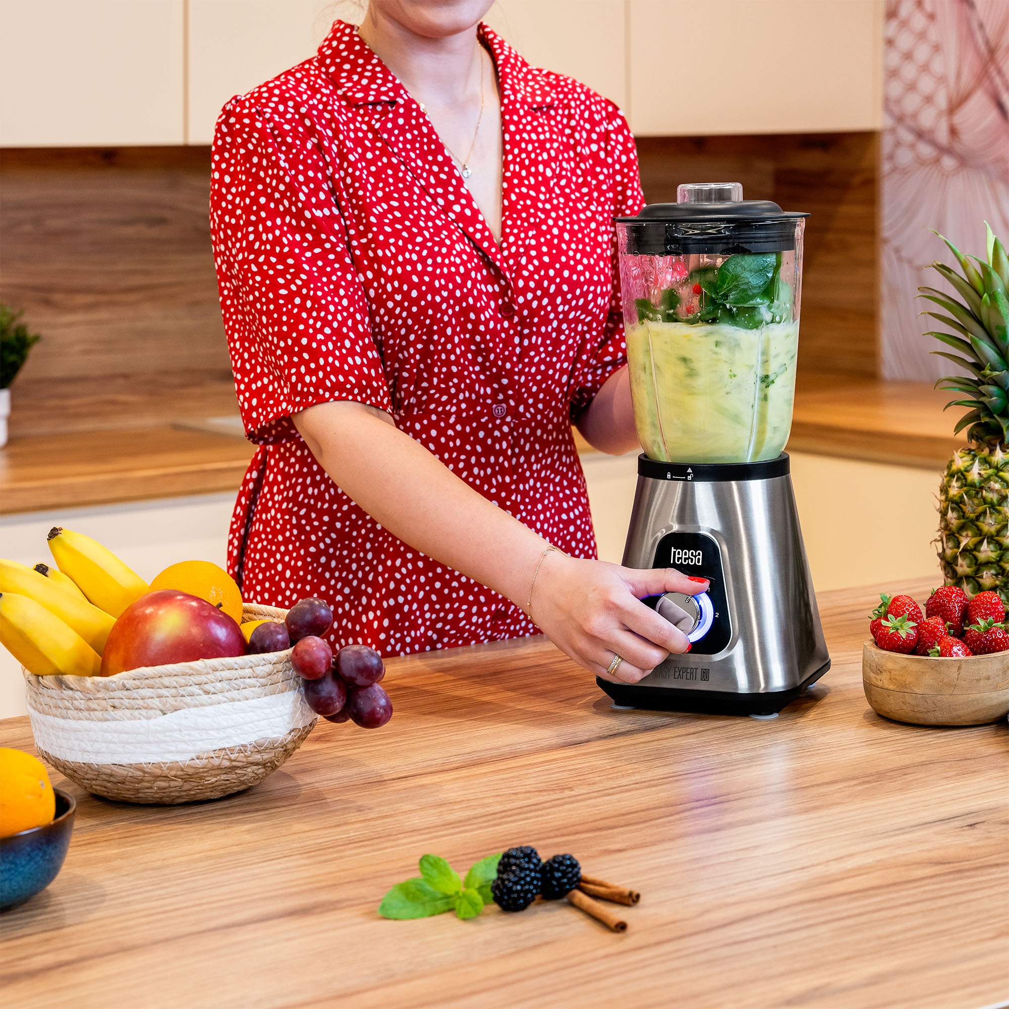 Kobieta w czerwonej sukni w grochy obsługuje srebrny blender z zielonym smoothie na drewnianym blacie.